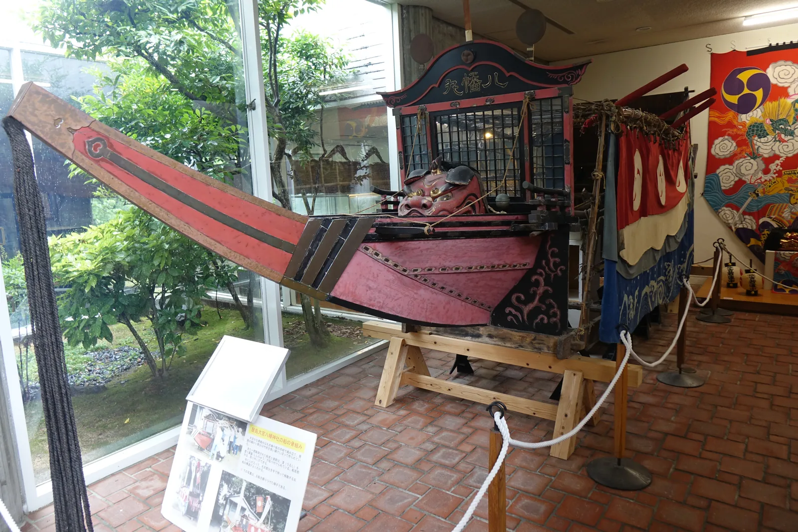 祭禮的展示室，前景是一座紅黑相間、船形的祭禮花車，其上有獅子頭，左側與後方是整面玻璃，玻璃外是中庭的樹木，右側掛著色彩鮮豔的大旗，地坪是紅磚
