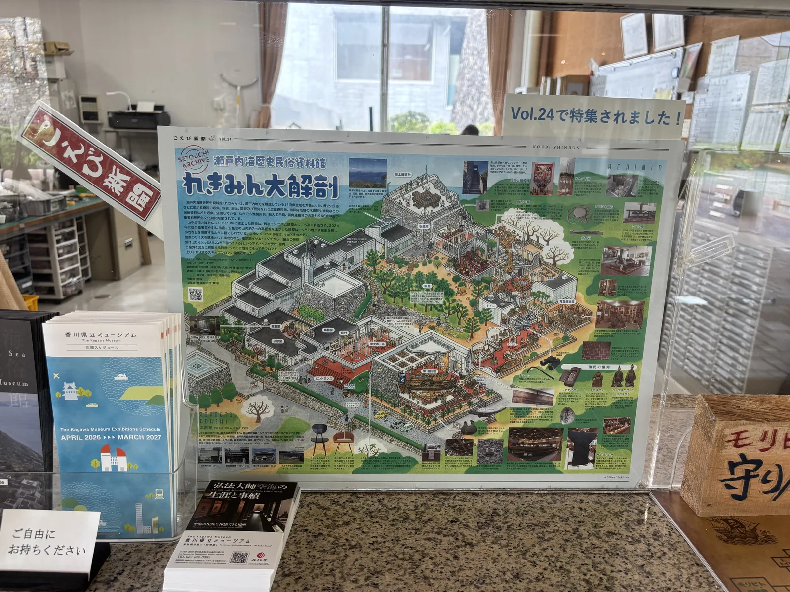 入口大廳的受付櫃台，櫃台上立著一塊壓克力板，內夾一張彩色的資料館鳥瞰插畫，旁邊放著摺頁與小冊子，背景是櫃台內的辦公空間與窗