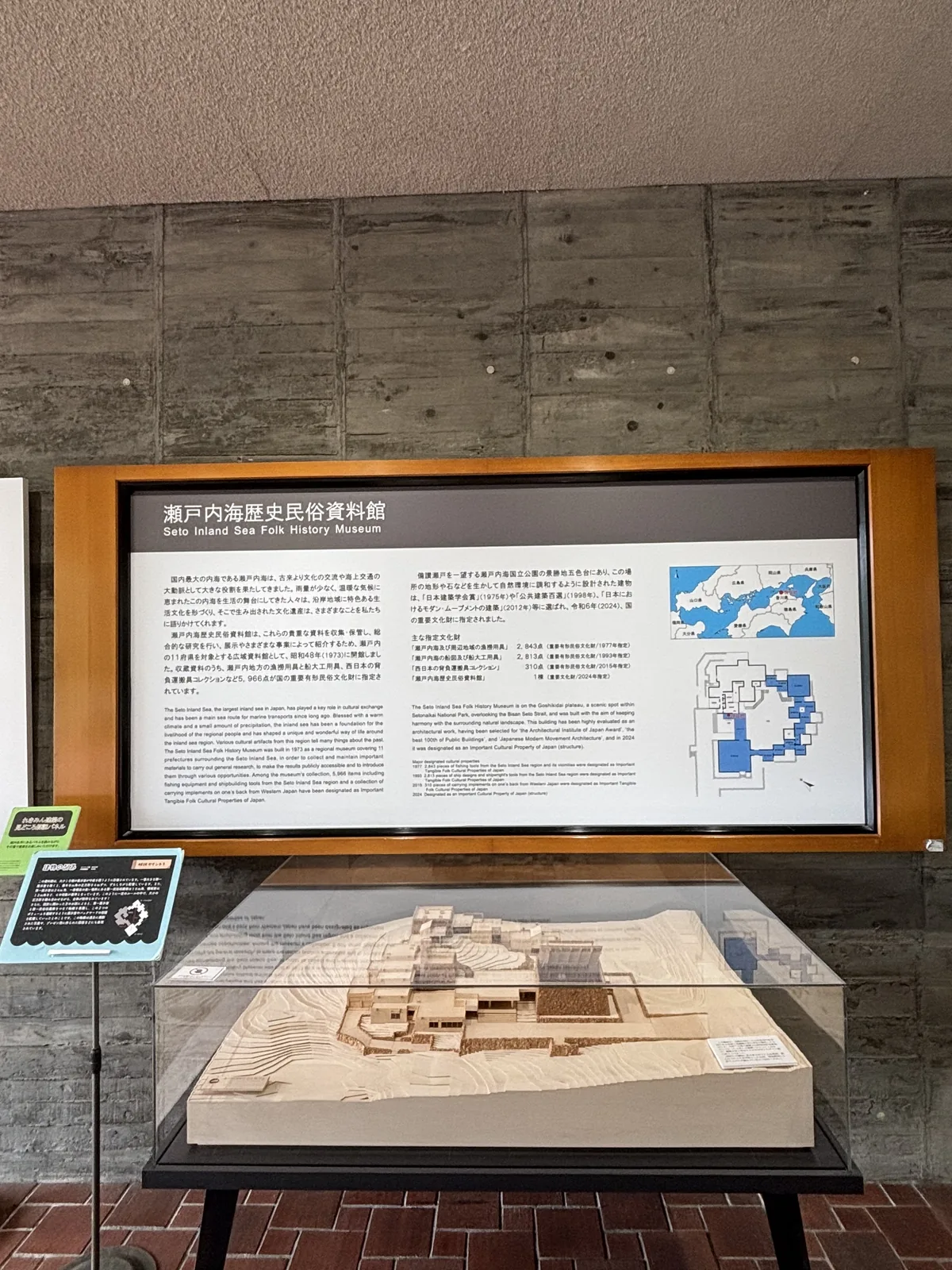 入口大廳一角，清水混凝土牆前立著一座木框的大型解說板，下方的玻璃罩內是資料館的淺色建築模型，模型呈現圍繞中庭、高低錯落的館舍，左側立著一塊小型的說明牌