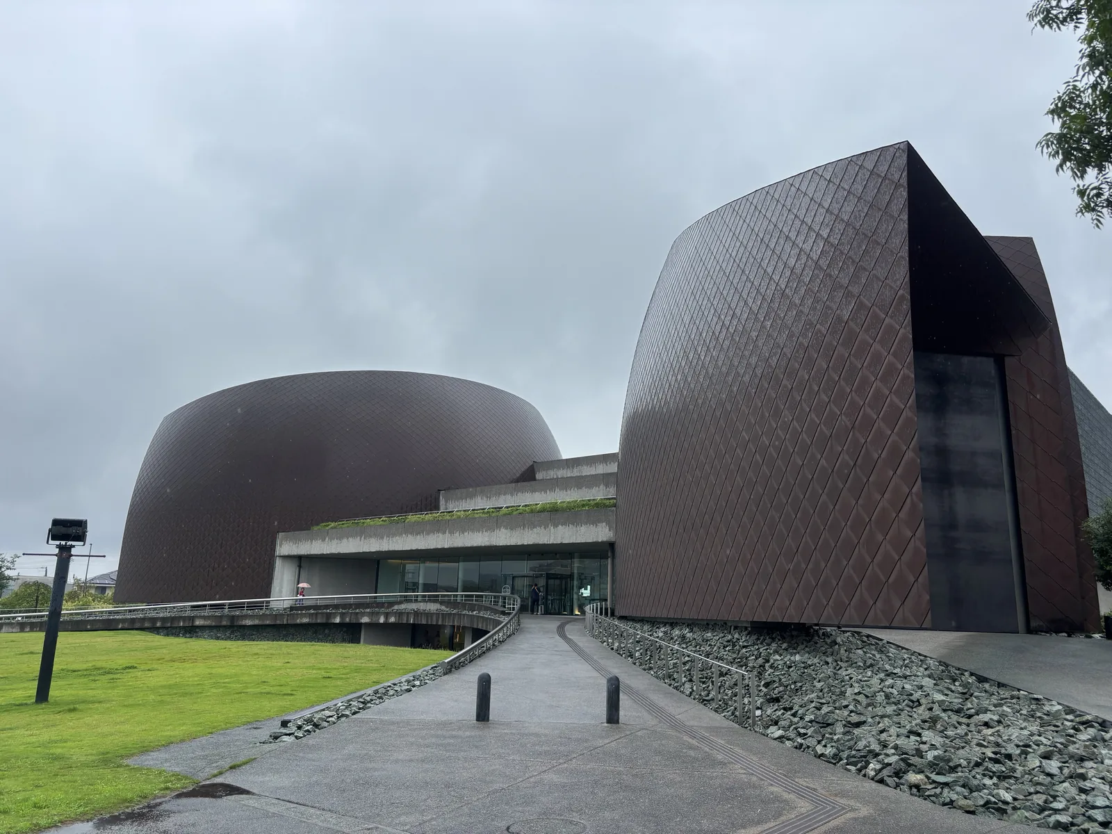 雨天的 Akagane Museum 外觀，前景是草坪與一條向前延伸的坡道，左側是一座低伏的圓鼓形銅板量體，右側是一座尖角高聳的銅板量體，表面為菱形紋樣，兩者之間是一層屋頂綠化的水平混凝土量體與玻璃入口，坡道右側鋪著灰綠色的碎石