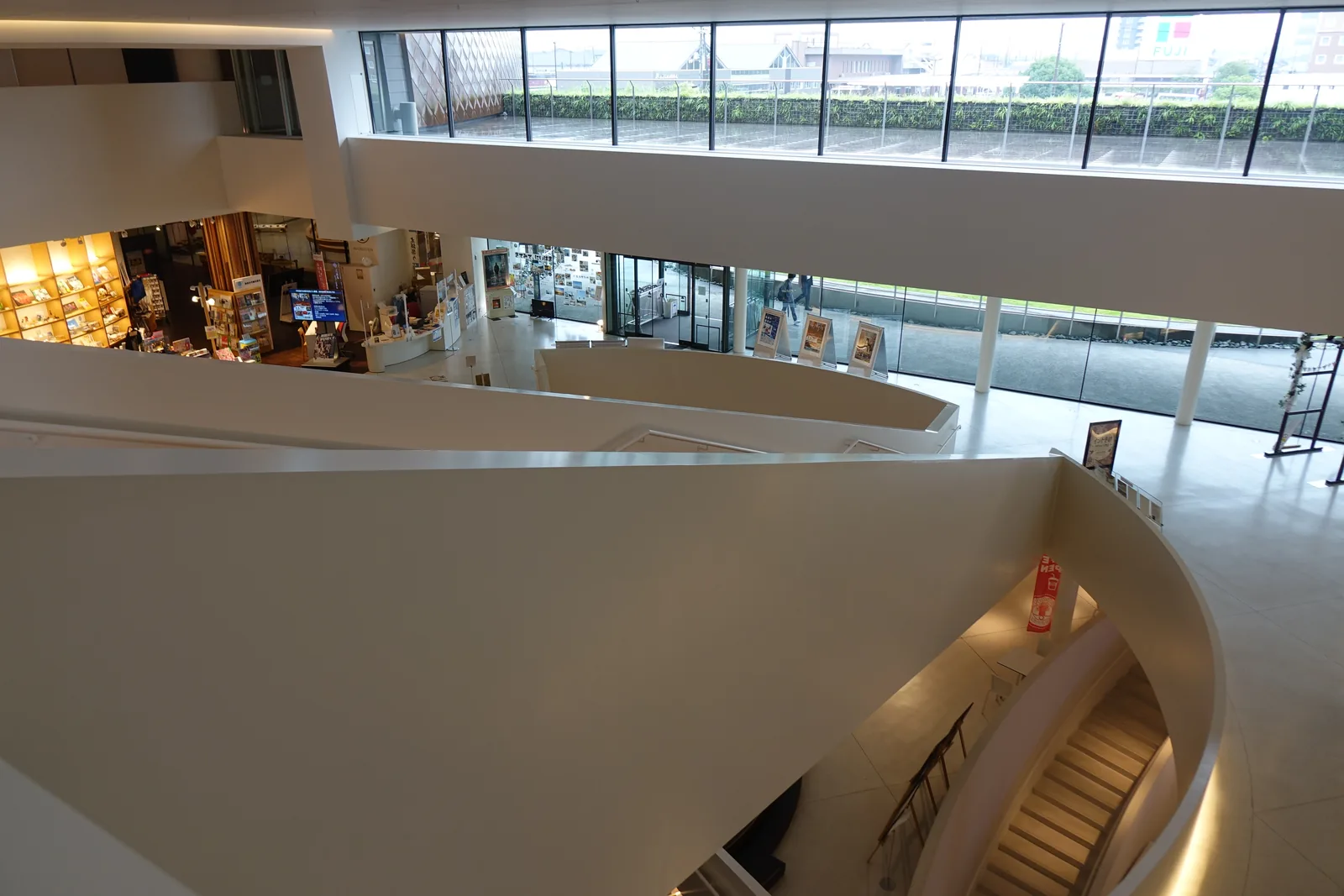 自上層俯視挑空，下方是入口樓層的商店、玻璃面與排列的展示板，中央是弧形的白色扶手牆與向下的樓梯，上方是一道橫向延伸的帶狀窗，窗外是屋頂綠化與市街