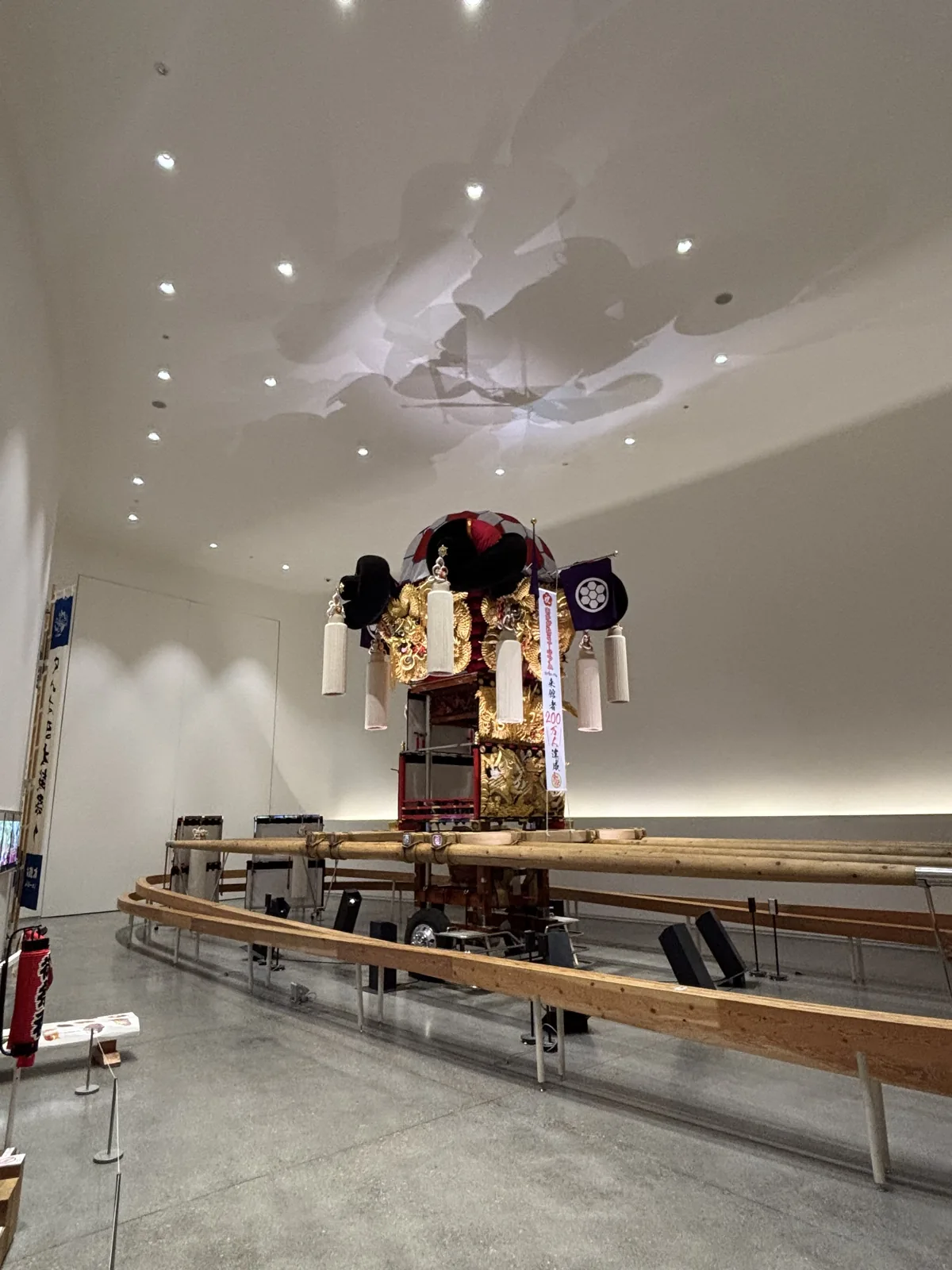 太鼓台 Museum，白色大室的中央展示著一座裝飾華麗的太鼓台，頂部有黑色與紅色的蒲團與白色流蘇，下方架著長木棒，周圍是一圈木製扶手，天花是白色曲面並投有雲形的光影