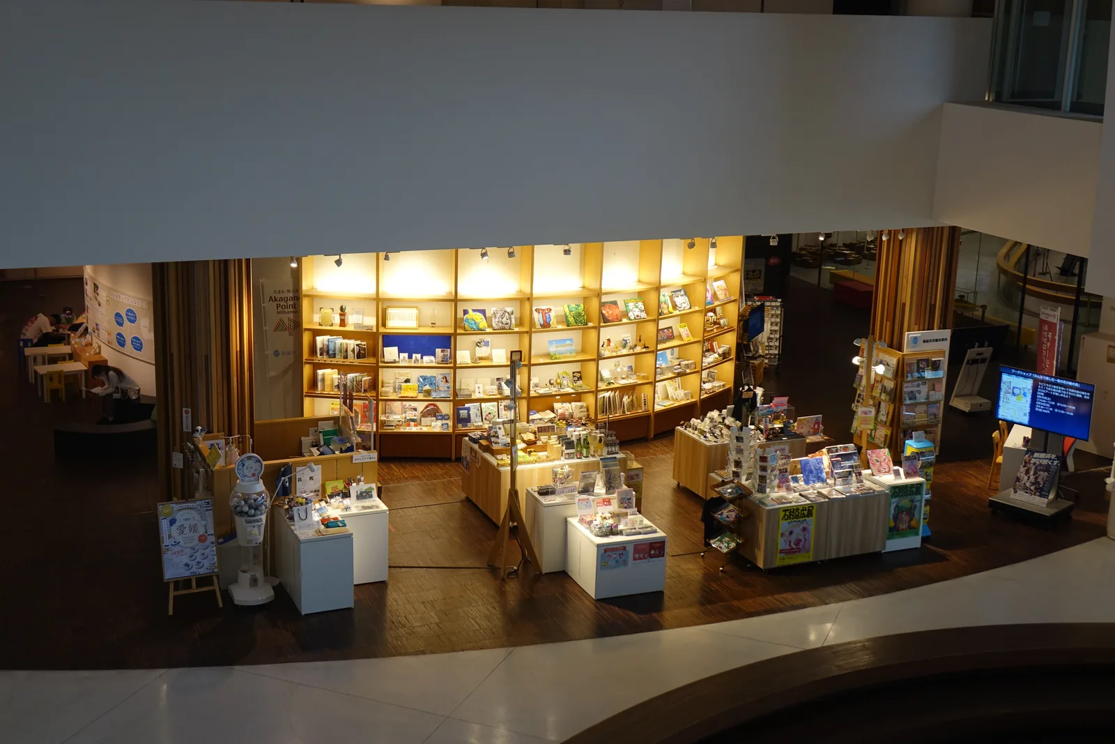 自上層俯視 museum shop，暖色照明的木製層架沿牆排列，擺滿書籍與商品，中央是幾座白色與木製的陳列台，左側有座椅，前景是白色的扶手牆