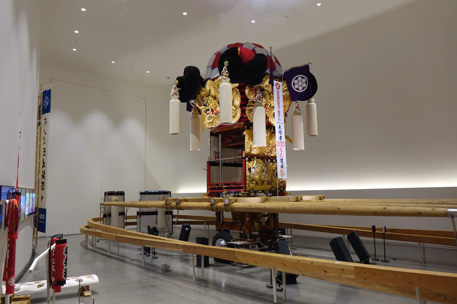 太鼓台 Museum 的另一角度，白色大室中央是一座高大的太鼓台，四周掛著白色的流蘇，周圍是一圈木製扶手，左側牆上掛著紅色的法被與長旗，天花是白色並嵌有燈