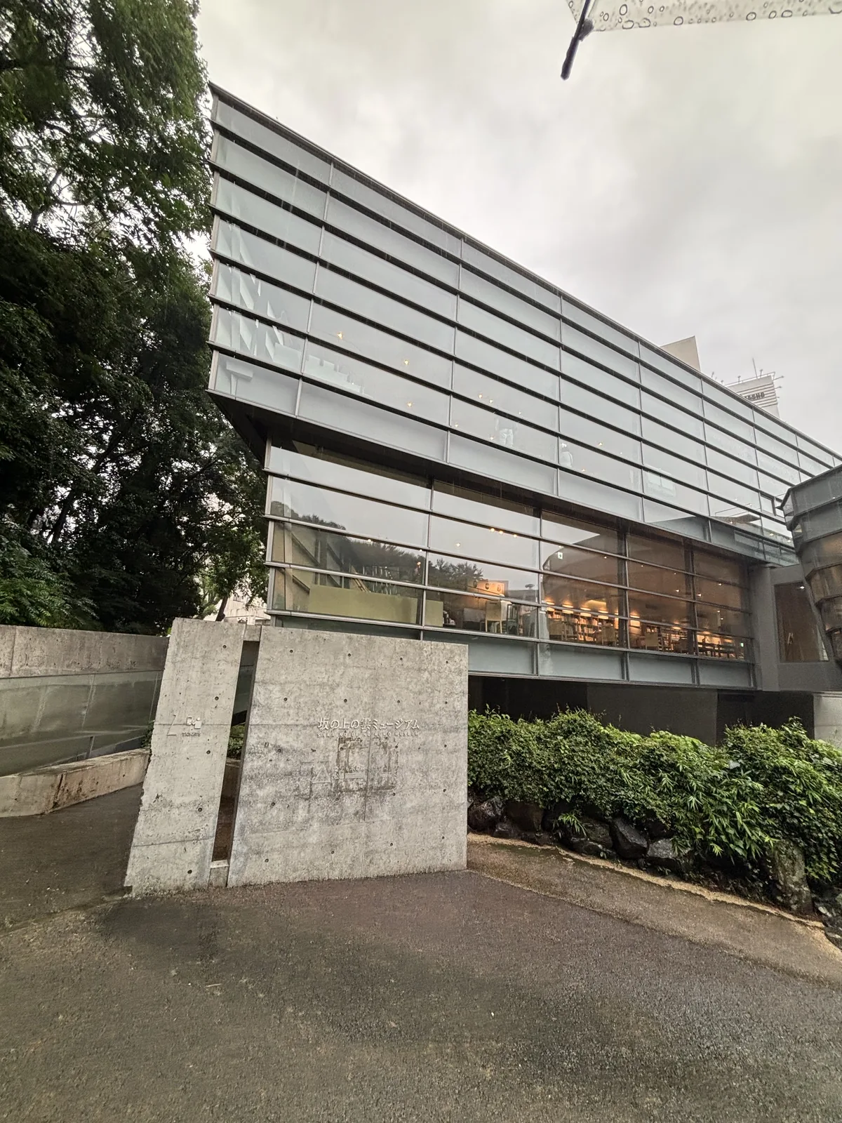 坂上之雲 Museum 的外觀，前景是一道清水混凝土的獨立牆與綠籬，後方是以水平帶分割的傾斜玻璃量體，玻璃面內可見暖色照明的室內，左側是大樹與城山的綠