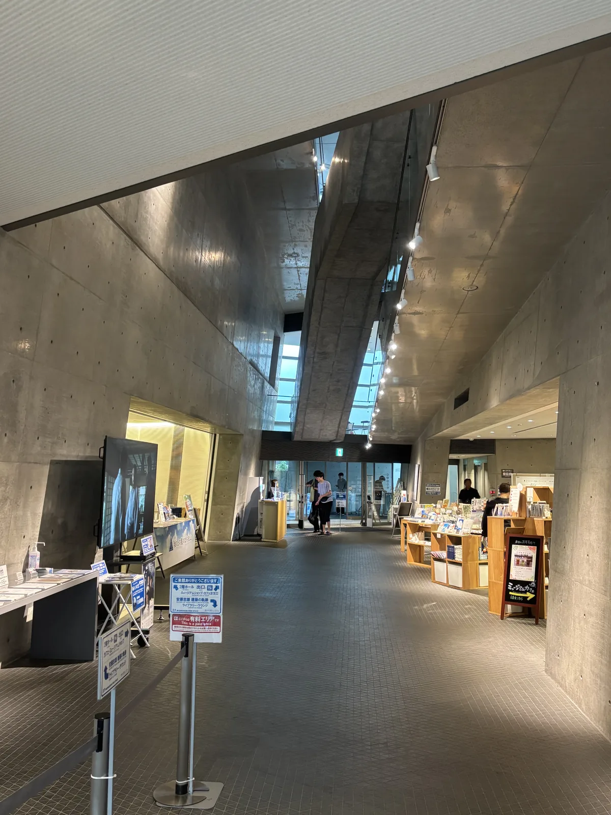 Museum 二樓大廳，兩側是清水混凝土的牆，上方是一道斜向穿過的混凝土量體與帶狀的照明，地面是深色的磚，中央排列著陳列台與書架，遠處是玻璃面與人影