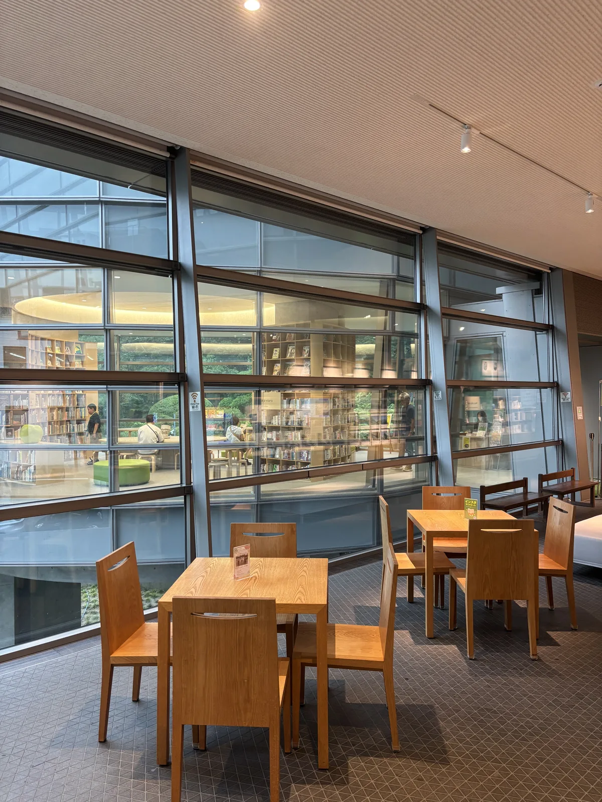 Library lounge 的木製桌椅，後方是一道以金屬框分割的玻璃壁，玻璃另一側可見兒童之書森林的弧形書架、閱覽的人與綠色坐墩，上方是帶狀的照明與玻璃外的綠
