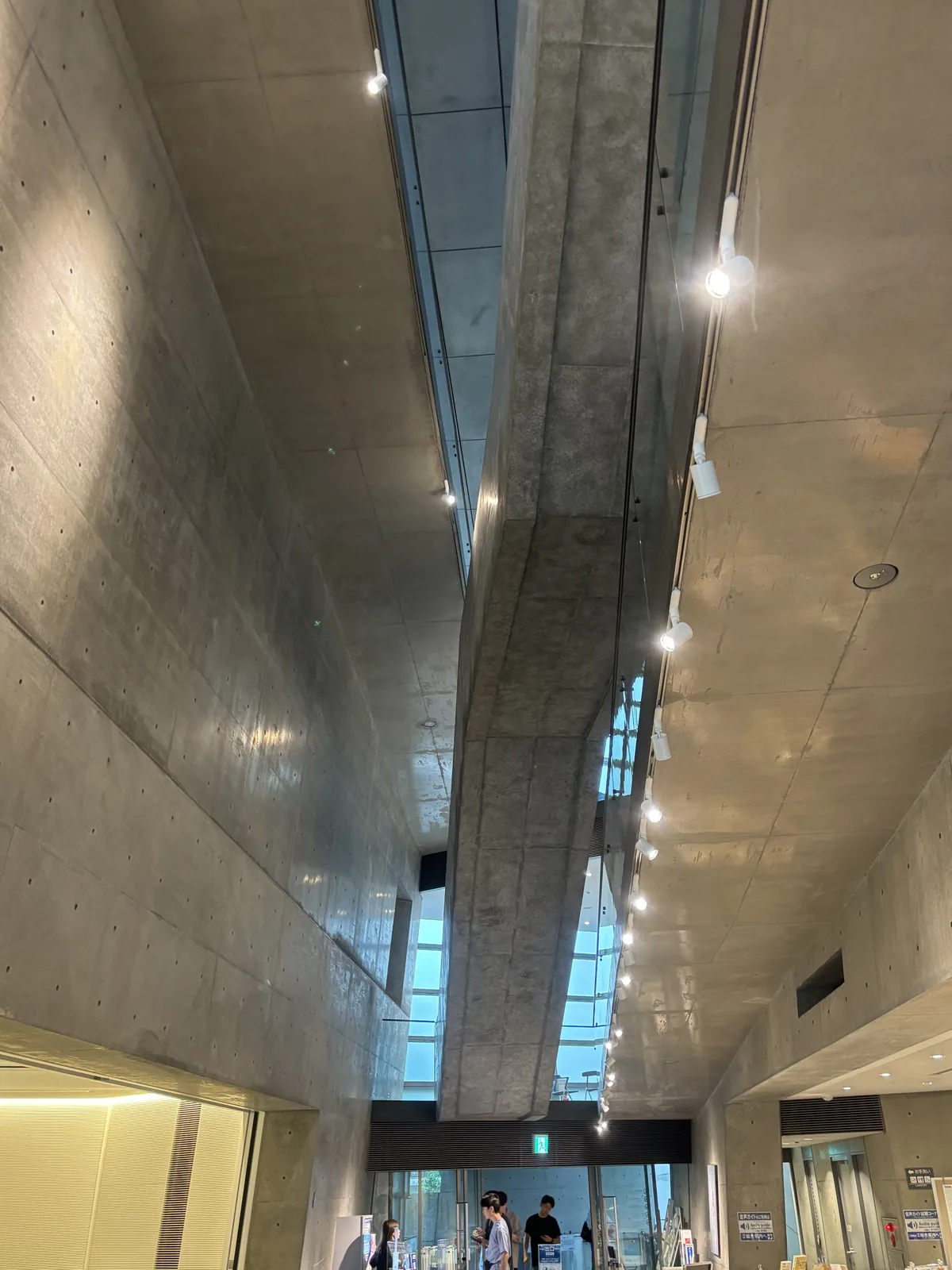 自下方仰看 Museum 挑空中斜跨的混凝土樓梯，樓梯底面是一道細長的混凝土板，斜向伸向上方，兩側是混凝土牆與玻璃，牆上排列著投射燈，下方可見參觀者與出入口