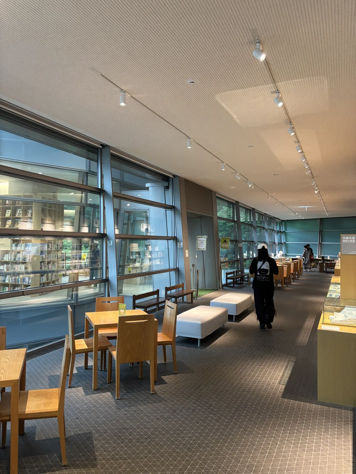 Library lounge 的木製桌椅與白色的長椅，左側是金屬框分割的玻璃壁，玻璃後是閱覽室的書架，右側是展示櫃，遠處有一名參觀者走向深處，天花是淺色的吸音板與軌道燈