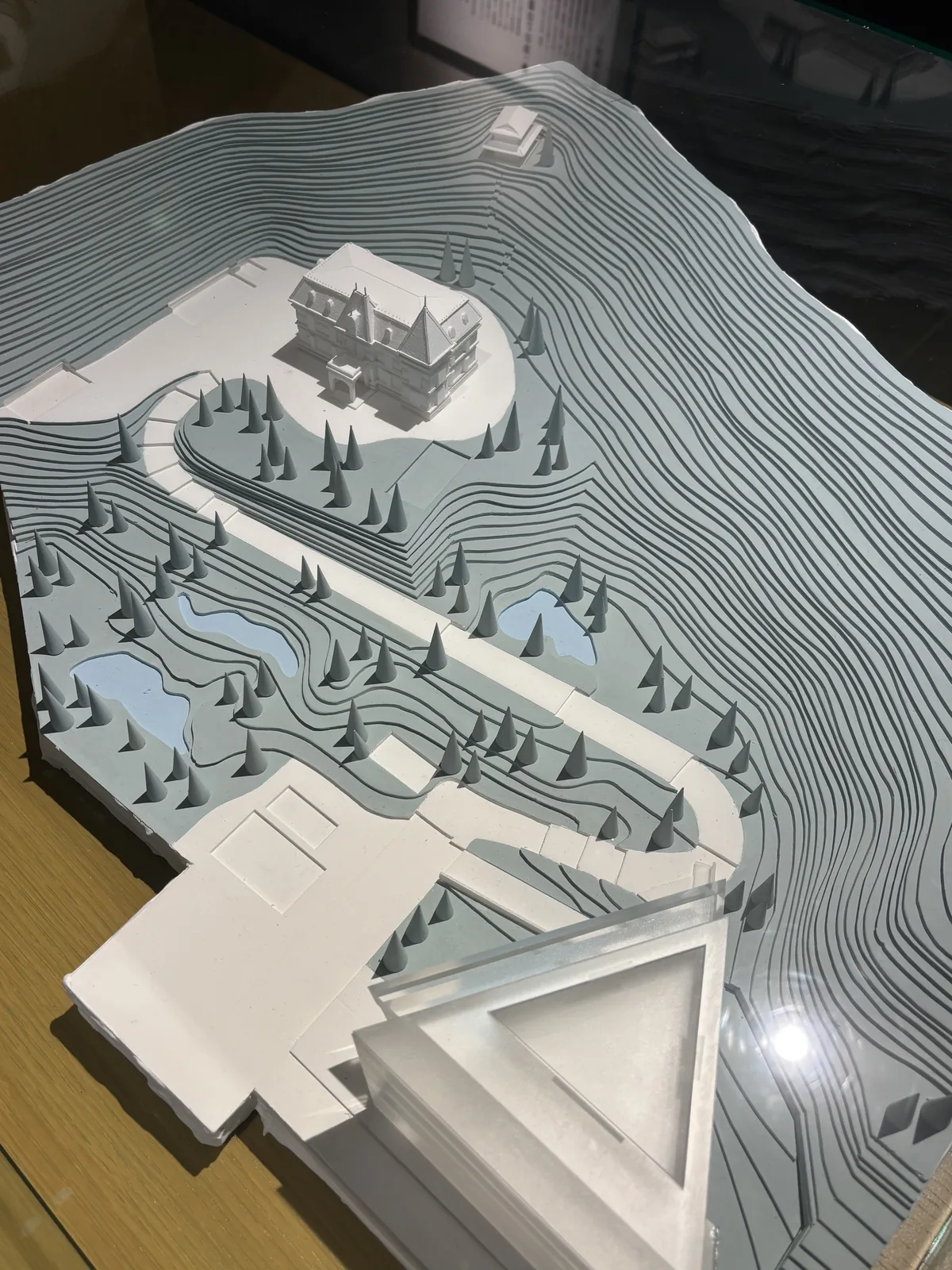 坂上之雲 Museum 與周邊的基地模型，白色的地形等高線上，上方是一棟法式洋館的模型，周圍是圓錐形的樹，下方是 Museum 的三角形量體與其旁的建築，一條彎曲的道路連接兩者