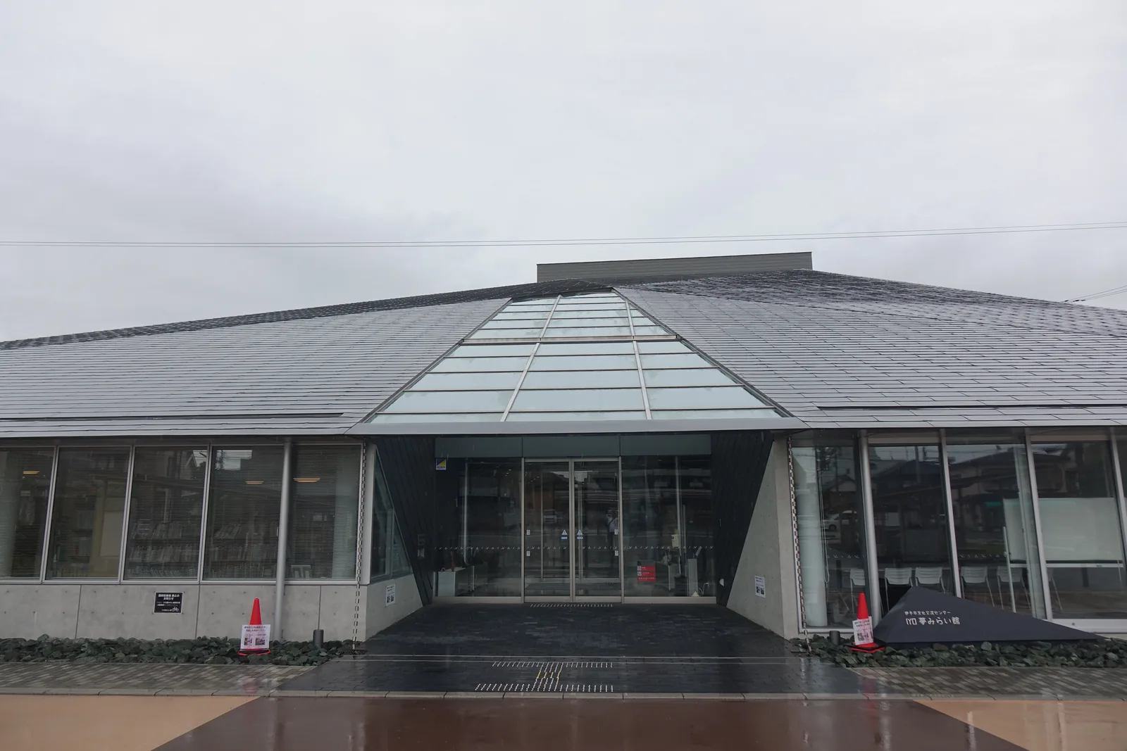 IYO夢未來館的正面入口，灰色的大屋頂自兩側向中央升起，中央是一道三角形的玻璃天窗，下方是玻璃的入口門與兩側的帶狀玻璃窗，窗內可見書架，入口前是濕潤的磚鋪廣場與碎石植栽帶，右側地面立著一塊黑色的三角形館名碑