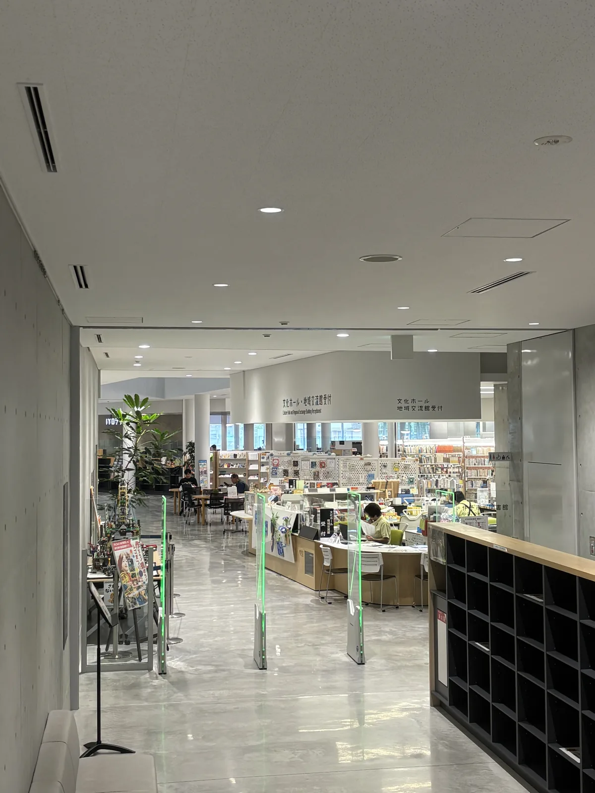 自入口通道望向館內，兩側是清水混凝土牆，右側是一排黑色的置物櫃，前方是 BDS 閘門的綠色燈框，閘門之後是挑高的一樓空間、服務台、書架與閱覽的人，地面是灰白色帶雲紋的光面地坪