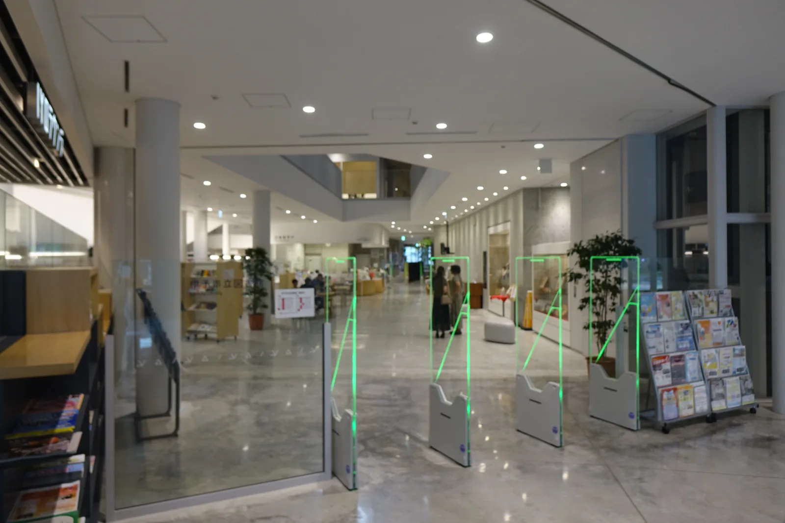一樓緣側 mall，前景是一排 BDS 閘門，左側是 IYO Lounge 的木格柵招牌與書架，中央是一根白色圓柱與盆栽，深處可見挑空、服務台與閱覽的人，地面是灰白色帶雲紋的光面地坪，右側是一座雜誌與傳單架