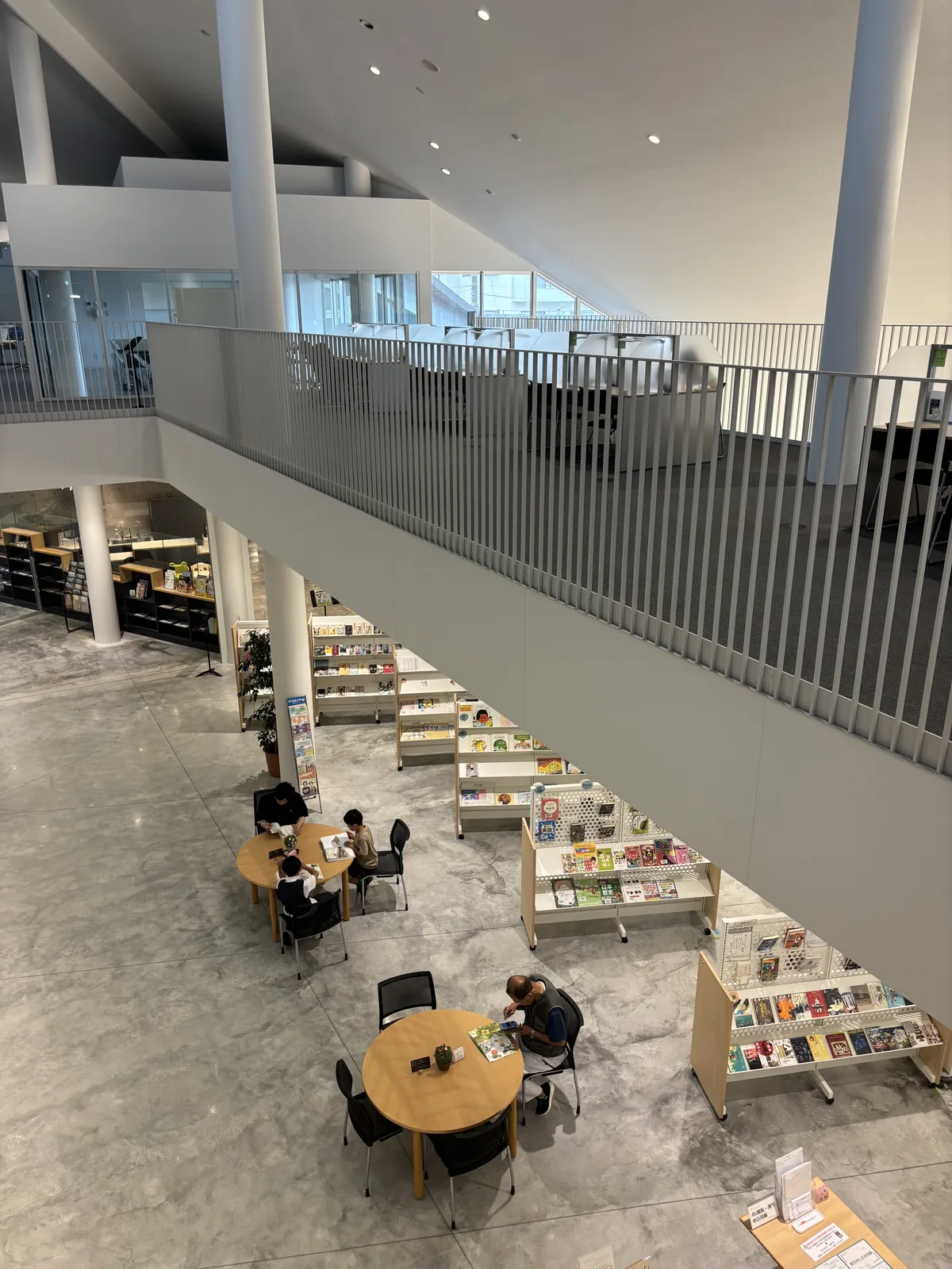 自二樓扶手俯看挑空，下方一樓是圓形木桌、黑色椅子與低矮的書架，地面是灰白色帶雲紋的光面地坪，對面是二樓的走道與白色直桿扶手，上方是兩根白色圓柱與斜向的天花