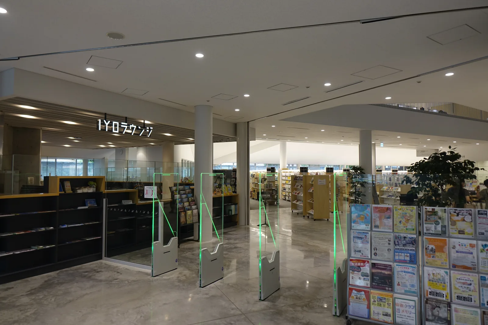 一樓的 IYO Lounge 入口，上方懸著白色的店名標示，下方是黑色的書架與木製的檯面，前方是一排 BDS 閘門，右側是白色圓柱、盆栽與傳單架，深處是一樓的開架書區與閱覽席