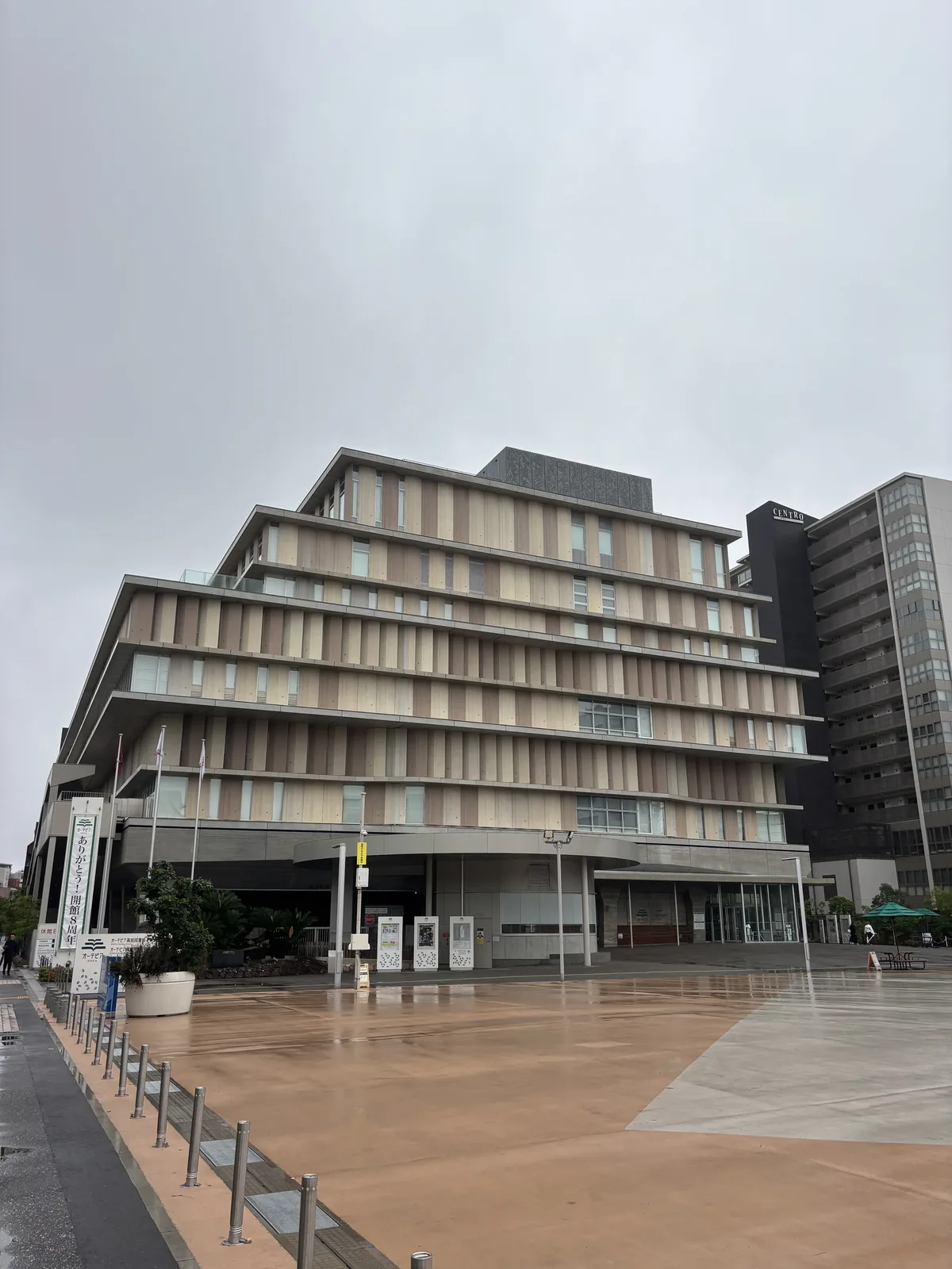 OTEPIA 西側的廣場與立面，前景是濕潤的淺褐色大面積地磚廣場，右側是一排低矮的金屬護柱，中景是多層的建築量體，每層樓板向外伸出薄的水平簷，簷間排列著淺色的垂直 louver，一樓退在深簷之下，右後方是一棟灰色的高樓