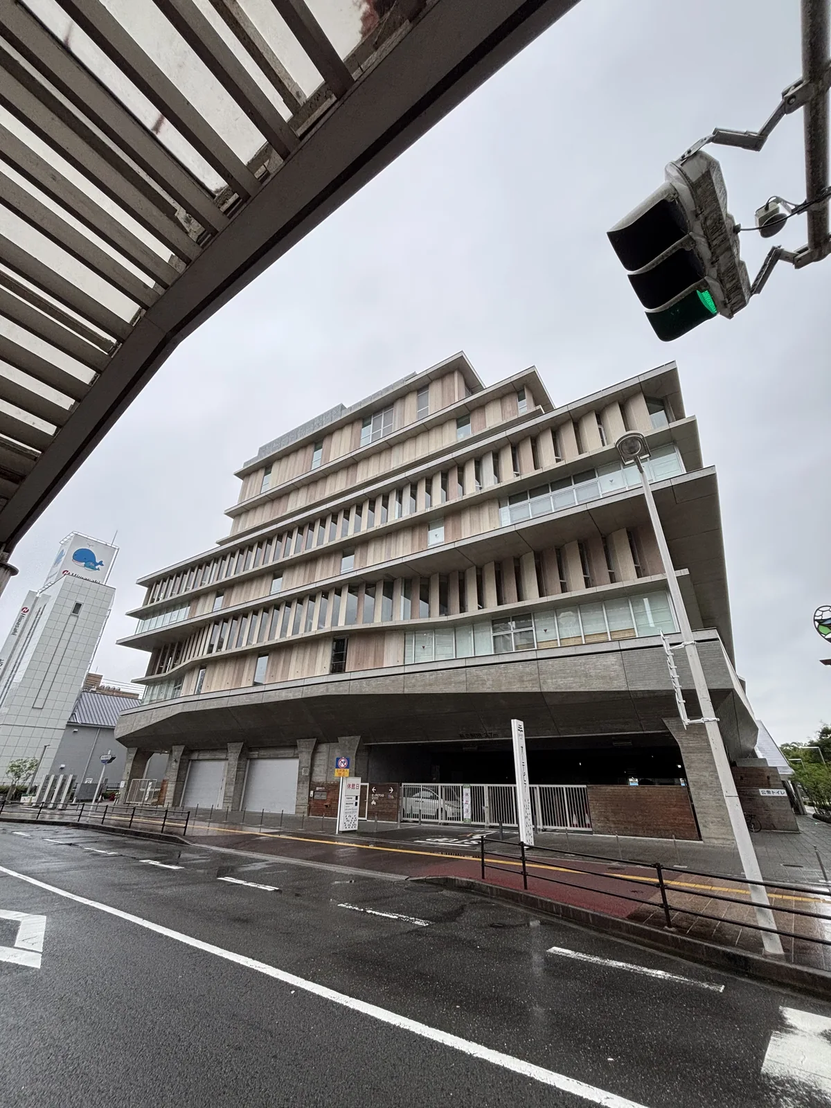 自東北側的街角仰看 OTEPIA，濕潤的柏油路面與斑馬線在前景，建築的東北角向上層層外伸出水平簷，簷間是淺色的垂直 louver，一樓是退縮的灰色基座與車庫入口，左上方是對街建築的格柵雨庇與一具紅綠燈，左遠方是一座白色的高塔
