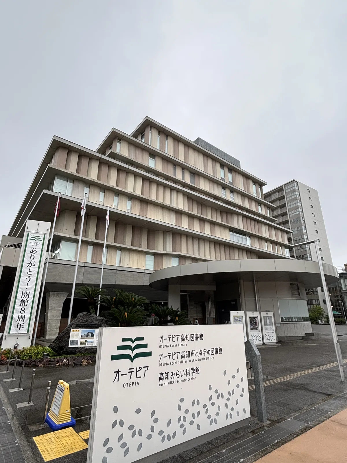 OTEPIA 西側入口前，前景是一塊白色的館名立牌，寫著三個館的名稱並印有綠色葉形標誌與散落的葉形圖案，立牌後方是入口前的植栽與一片圓弧形的雨庇，再後方是層層外伸的水平簷與淺色 louver，左側立著寫有開館八週年字樣的直幅布條與數根旗桿