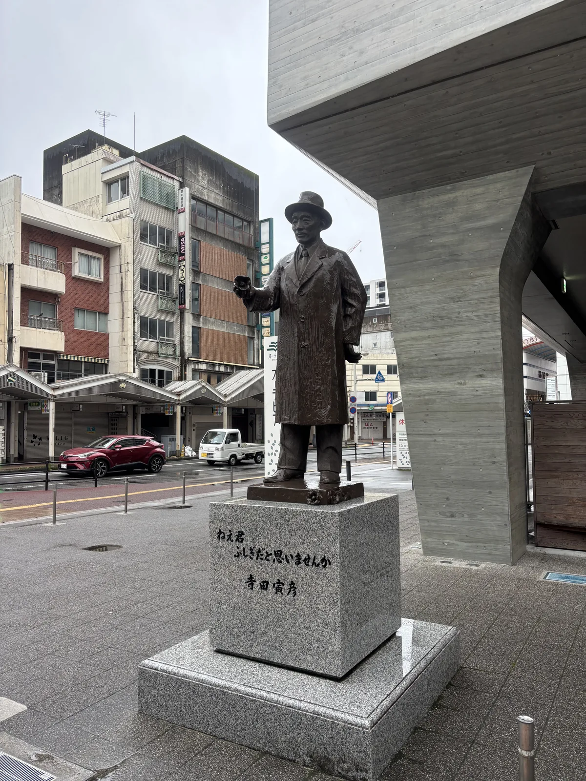 OTEPIA 東側懸挑的清水混凝土量體之下，立著寺田寅彥的全身銅像，銅像戴帽穿大衣、一手持帽，立在灰色花崗岩台座上，台座刻著一行文字與寅彥的名字，右側是一根表面帶有木紋模板痕跡的混凝土柱，背景是街道對面的商店拱廊與住商建築