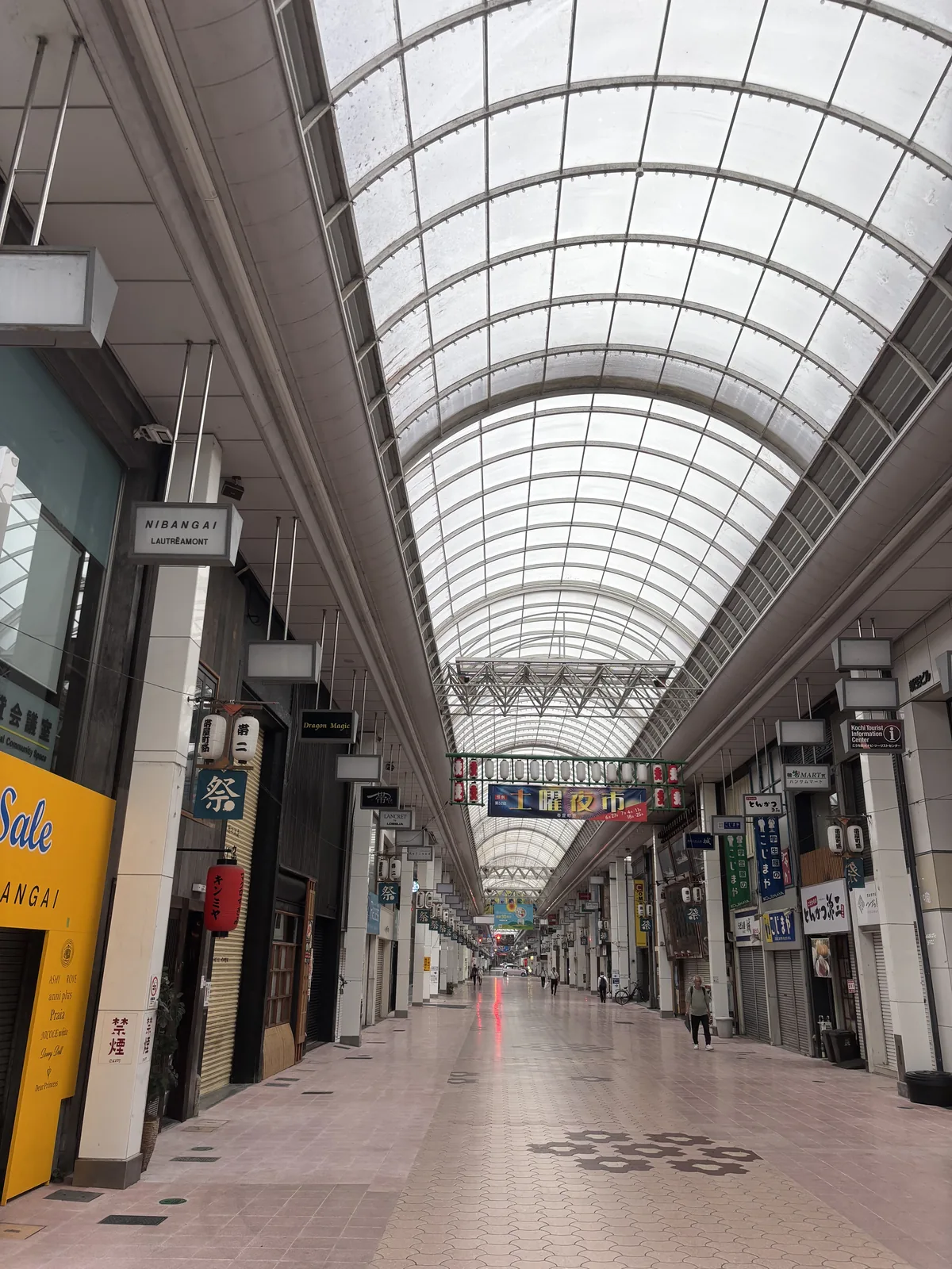 帶屋町商店街的拱廊，上方是半透明的拱形屋頂與白色的鋼架，兩側是拉下鐵捲門的商店與直立的招牌，地面鋪著淺粉色的地磚與圓形的圖案，深處拱廊一路延伸，有零星的行人