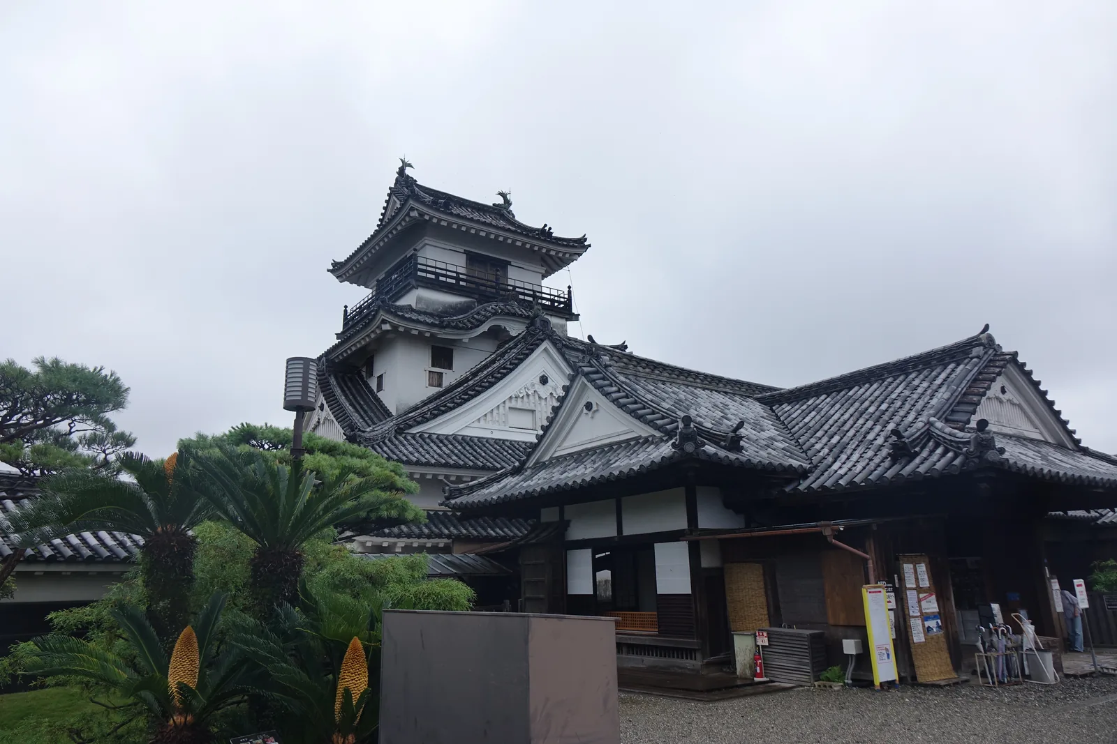 本丸上的懷德館與天守，左後方是白壁黑簷的天守，右側是一棟一層的御殿建築，屋頂為大型的入母屋造本瓦葺，正面突出一座玄關的屋頂，前景是棕櫚與蘇鐵等植栽，右側有幾名參觀者站在入口前