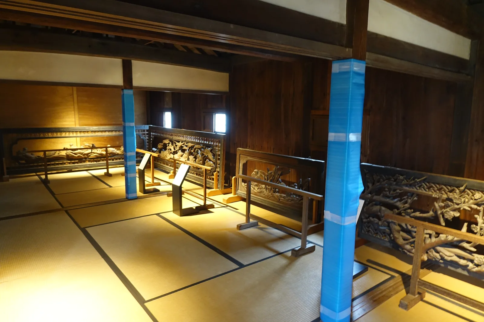 本丸建築內的展示室，榻榻米地板上沿牆排列著數件大型的木雕欄間，以木架支撐，兩根柱子以藍色的保護材包覆，牆面是深色的木板與白色的壁面，上方是木造的梁與小窗