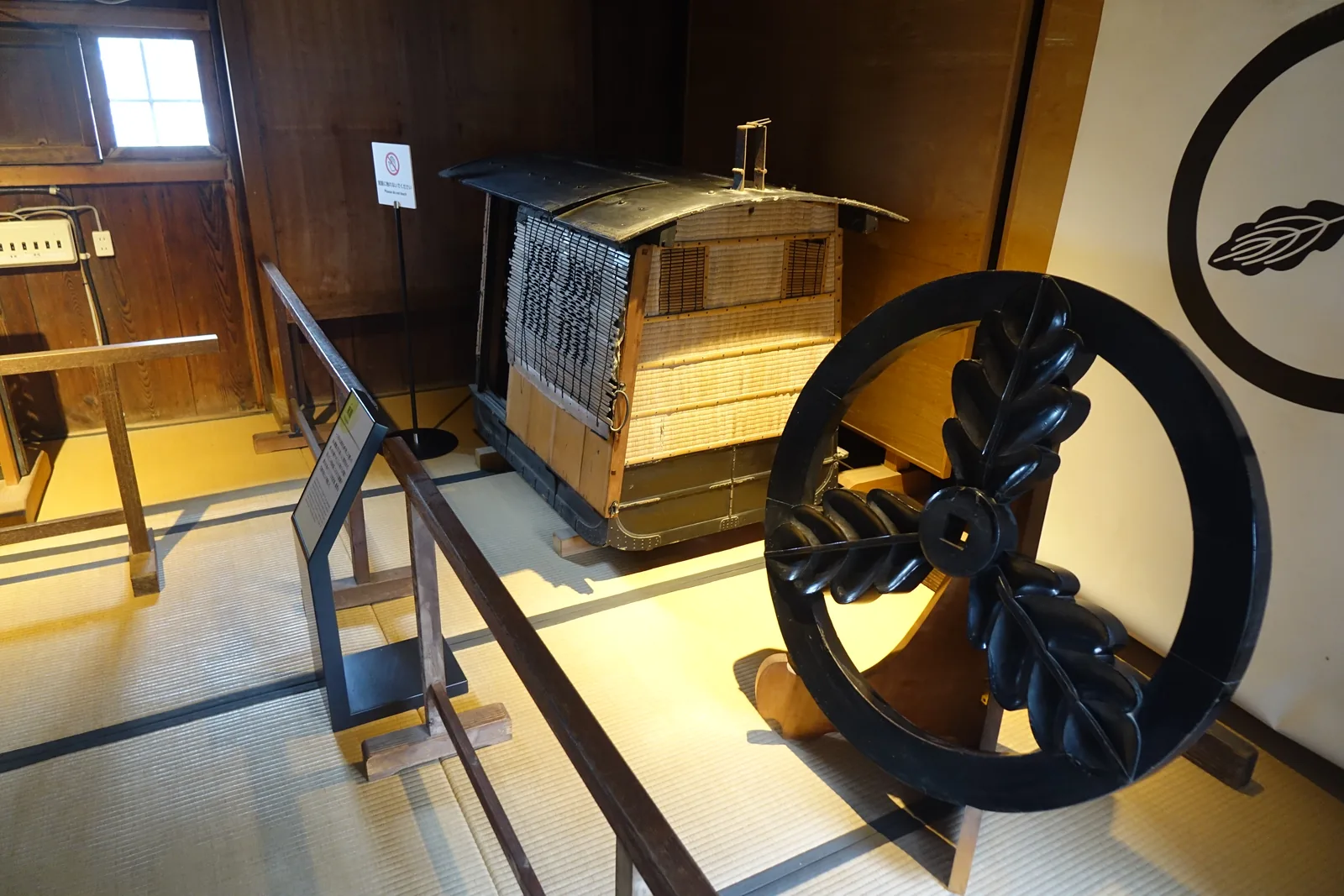 本丸建築內的展示室，榻榻米地板上放置著一頂木造的駕籠，前方是一座黑色的大型家紋圓輪，牆面是深色的木板與白色的壁面，上方是一扇小窗與木梁，四周有木製的柵欄與解說牌