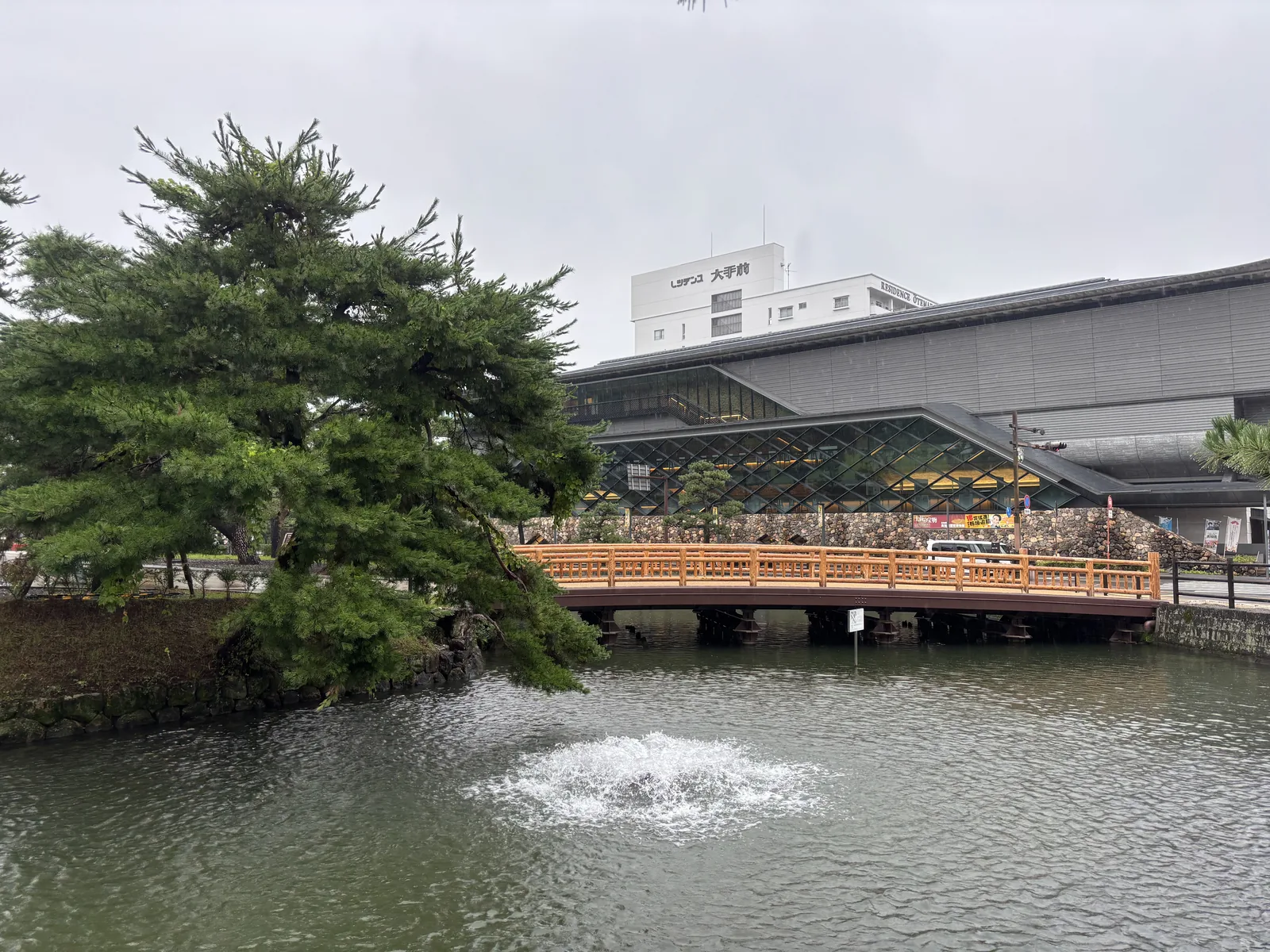 高知城前的護城河與朱色欄杆的木橋，水面中央有一處噴水，左側是一株枝葉濃密的松樹，橋的後方是博物館低平的量體，可見深出的屋簷、菱形格子的立面與石垣狀的基座，最上方是一棟白色的高樓