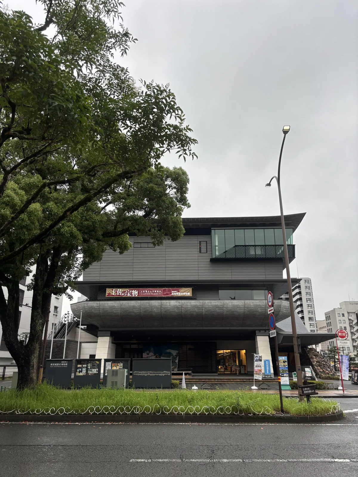 博物館北側面向追手筋的立面，二樓伸出一片船形的混凝土陽台，弧形的底面帶有木紋般的紋理，陽台上懸著展覽的布條，一樓退縮為 piloti，柱間是寬大的台階與玻璃入口，前景是街道與中央分隔帶的草地，左側立著館名的看板