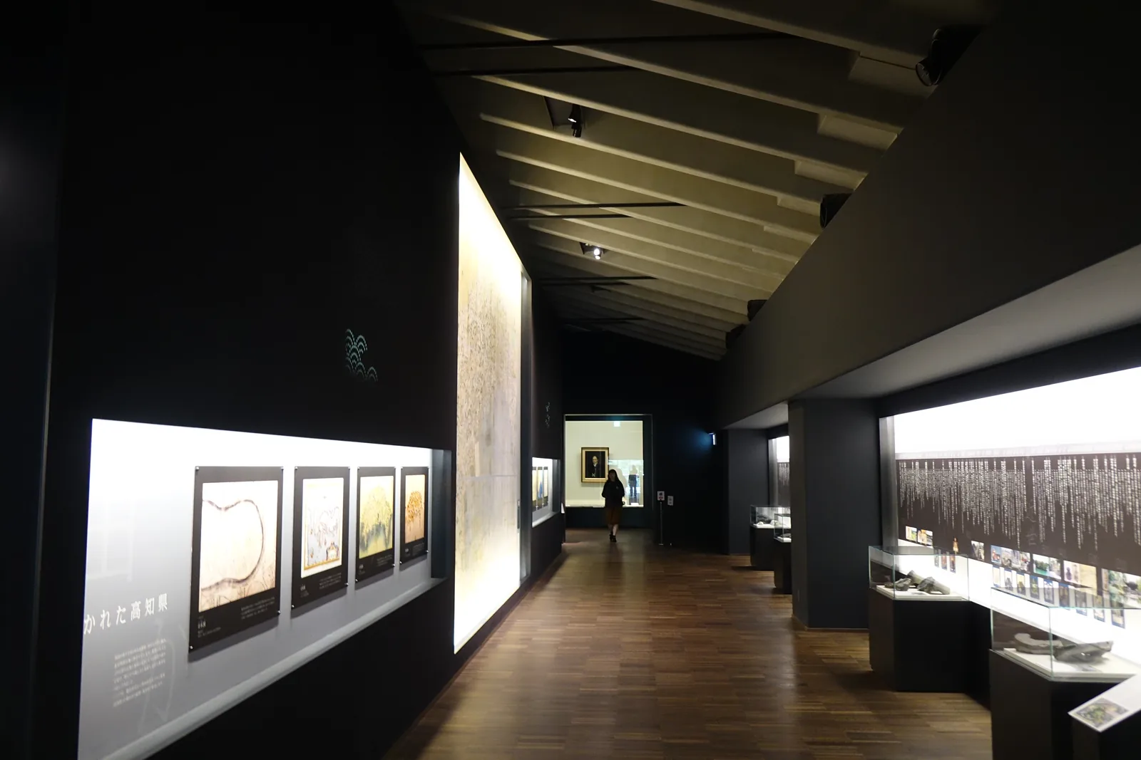 三樓展示室之間的長通路，左側是一道發光的白色大型壁面與幾幅框裝的地圖，右側是低矮的展示櫃與深色的牆面，天花的梁沿通路方向排列，地面是深色的木地板，盡頭是一幅明亮的肖像與一名參觀者