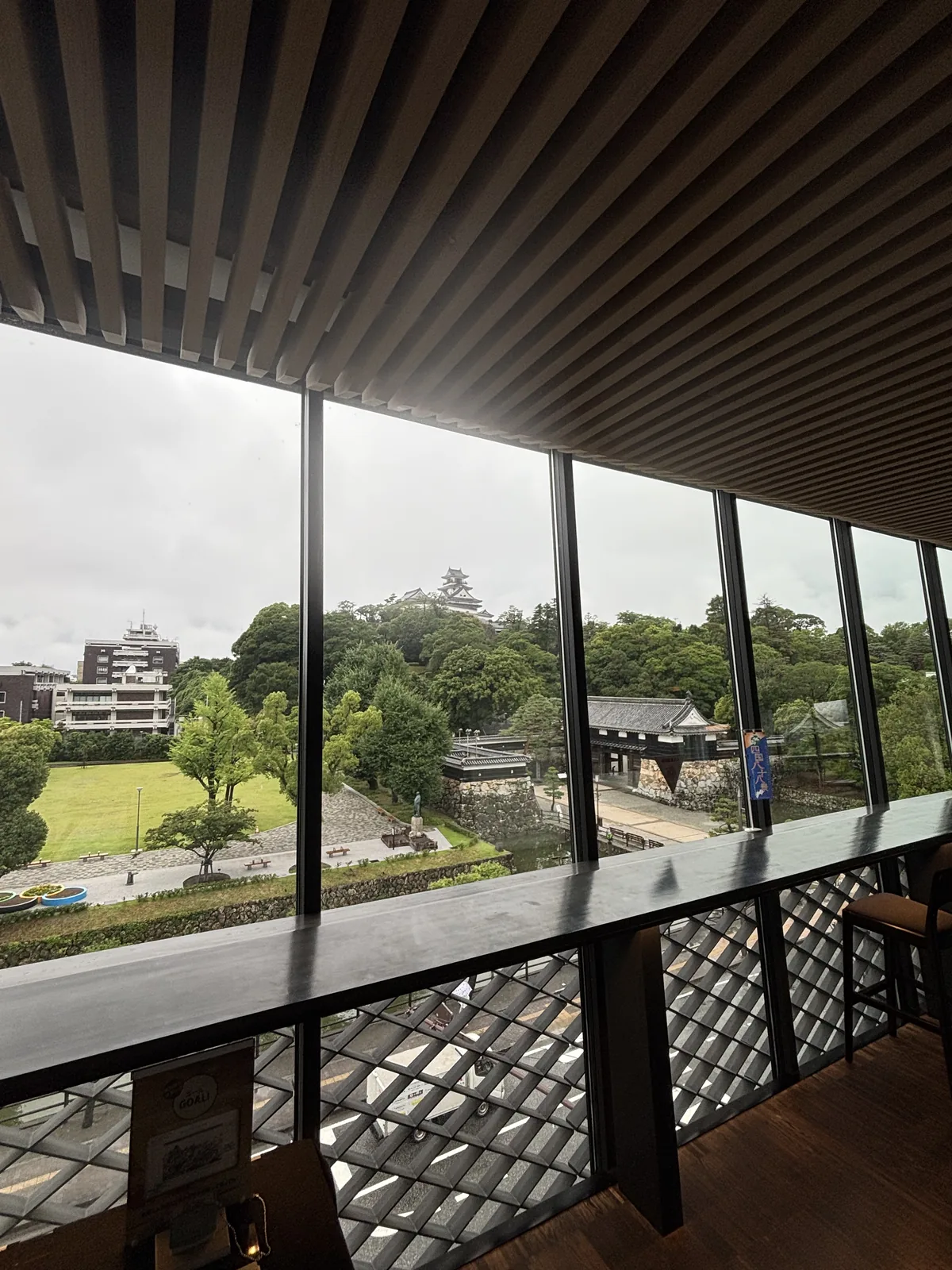 三樓的高知城展望 lobby，上方是成排的檜木 louver，前方是一整面以細框分割的落地玻璃，窗前是一道長長的深色檯面，檯面下是菱形格子的欄杆，玻璃外是草地、追手門、樹林與城山上的天守