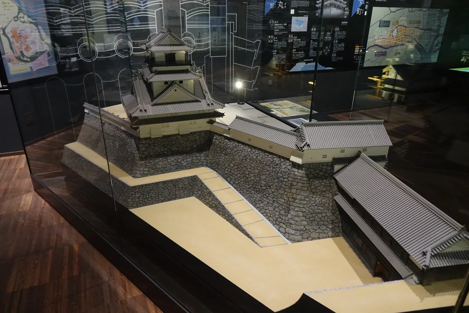 展示室內的城郭模型，玻璃櫃中是一座以灰色石垣、白色塀與黑瓦屋頂構成的城門與桝形模型，模型後方是深色的牆面與幾幅地圖、文字的展板，地面是深色的木地板