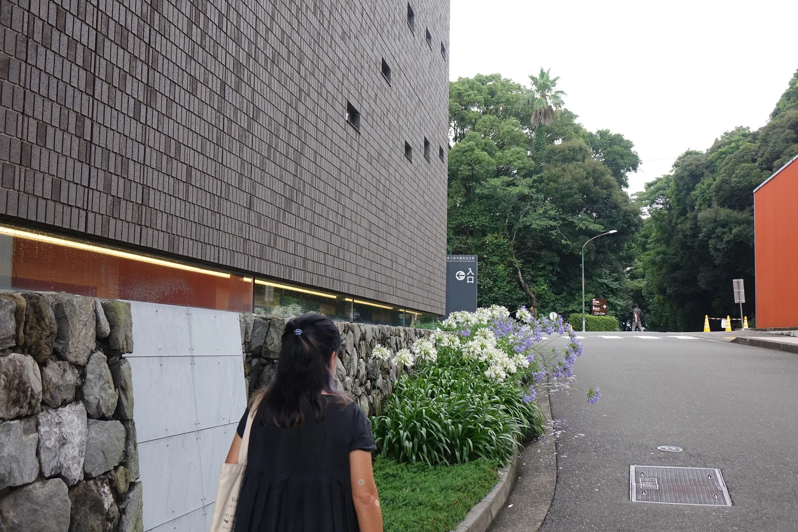 高知縣立坂本龍馬紀念館新館的北側立面，深褐色磁磚牆面上零星開著幾個小方窗，牆腳是石垣與白色的花叢，車道盡頭可見本館朱紅色的量體
