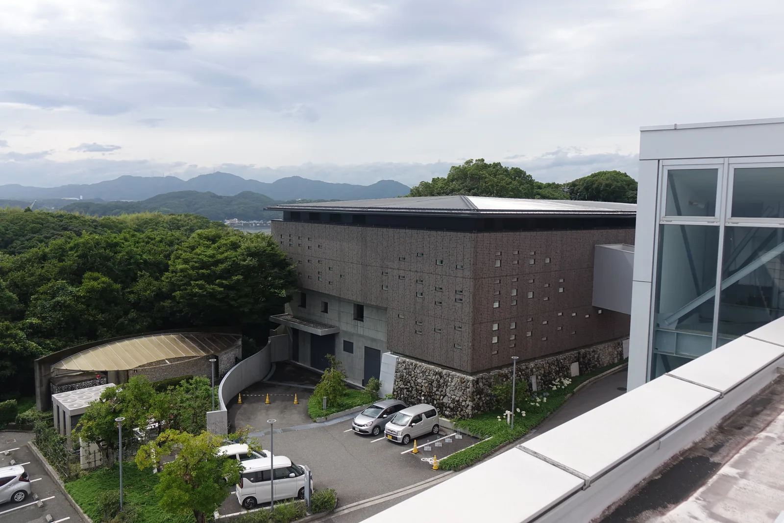 自本館屋頂俯瞰新館，深褐色磁磚包覆的方正量體上覆一片淺色的金屬屋頂，牆腳有一段石垣，前方是停車場與樹林，遠處是高知平野與山稜
