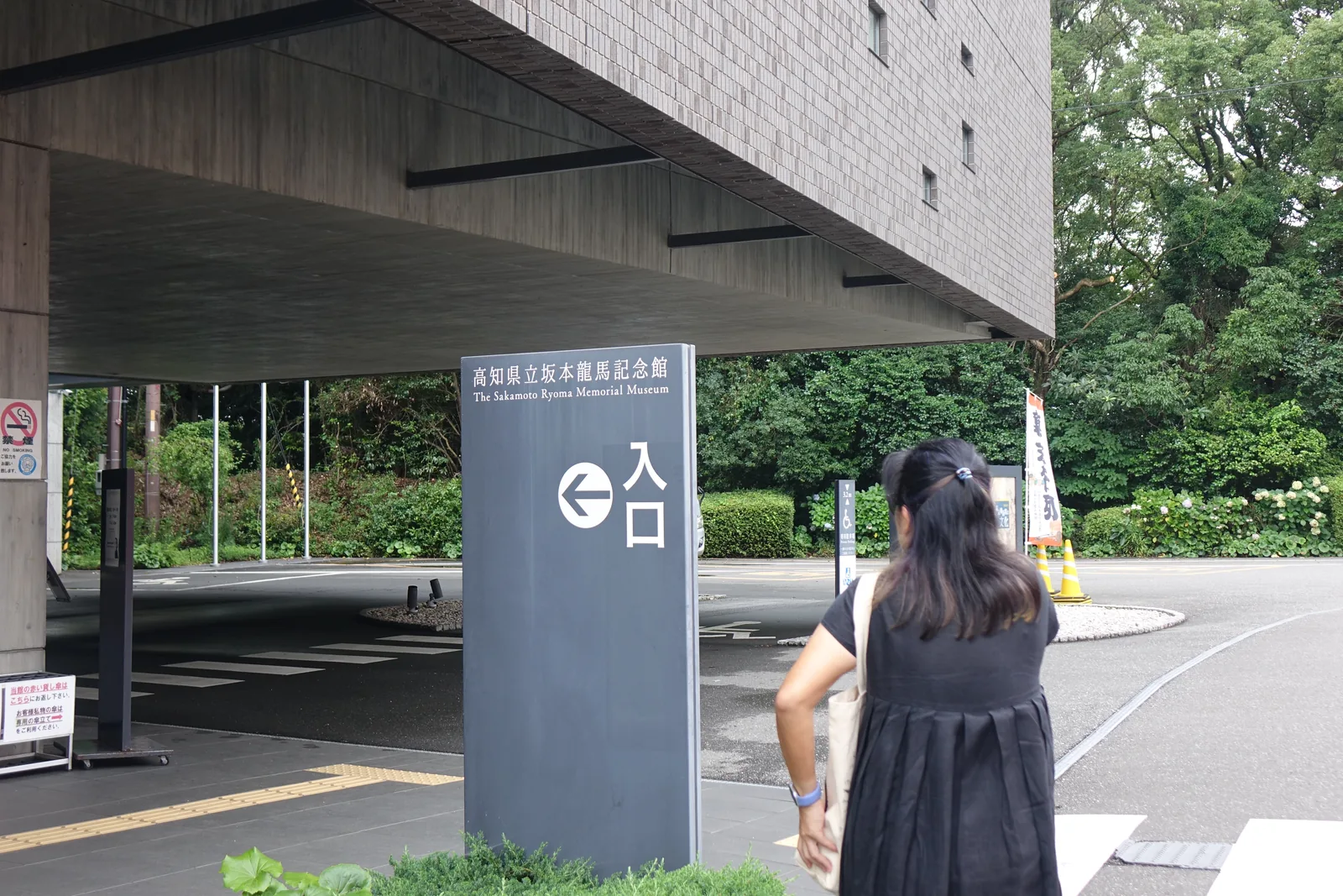 新館東側出挑量體之下的入口前，混凝土的挑簷底面與磁磚牆面在上方，前方立著一座深灰色的入口指標，遠處是車寄與樹林