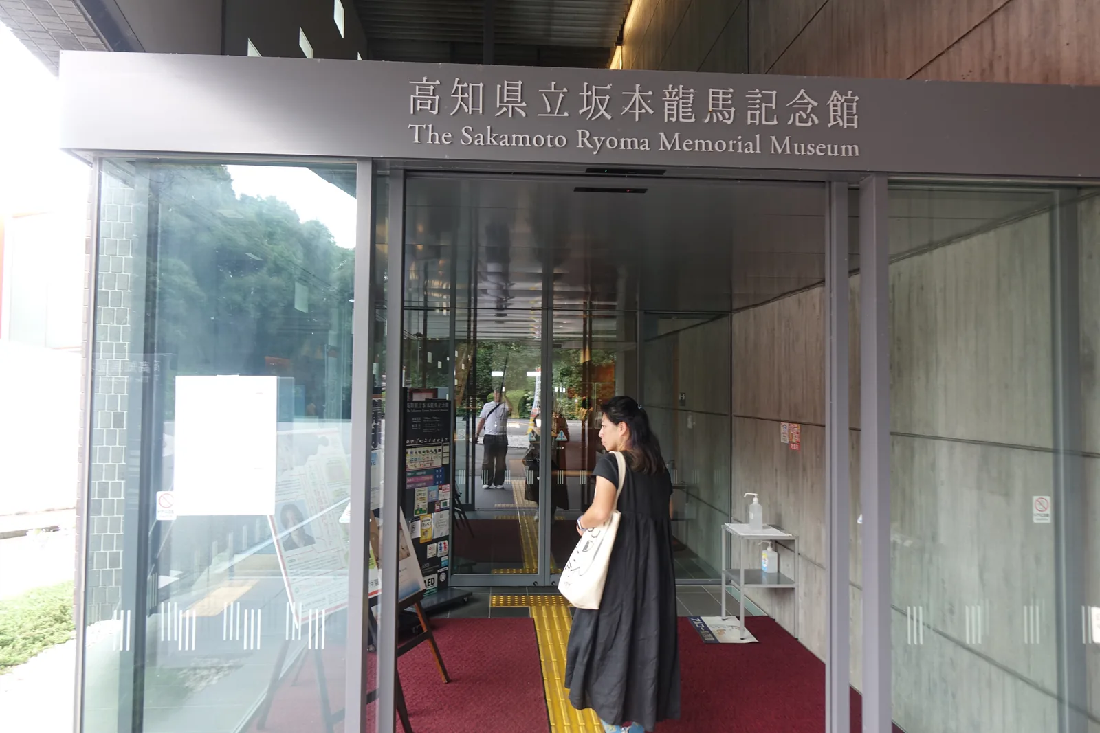 新館入口的玻璃門，上方掛著寫有館名與英文名的深灰色招牌，門內是紅色的地墊與導盲磚，兩側是木紋與混凝土的牆面