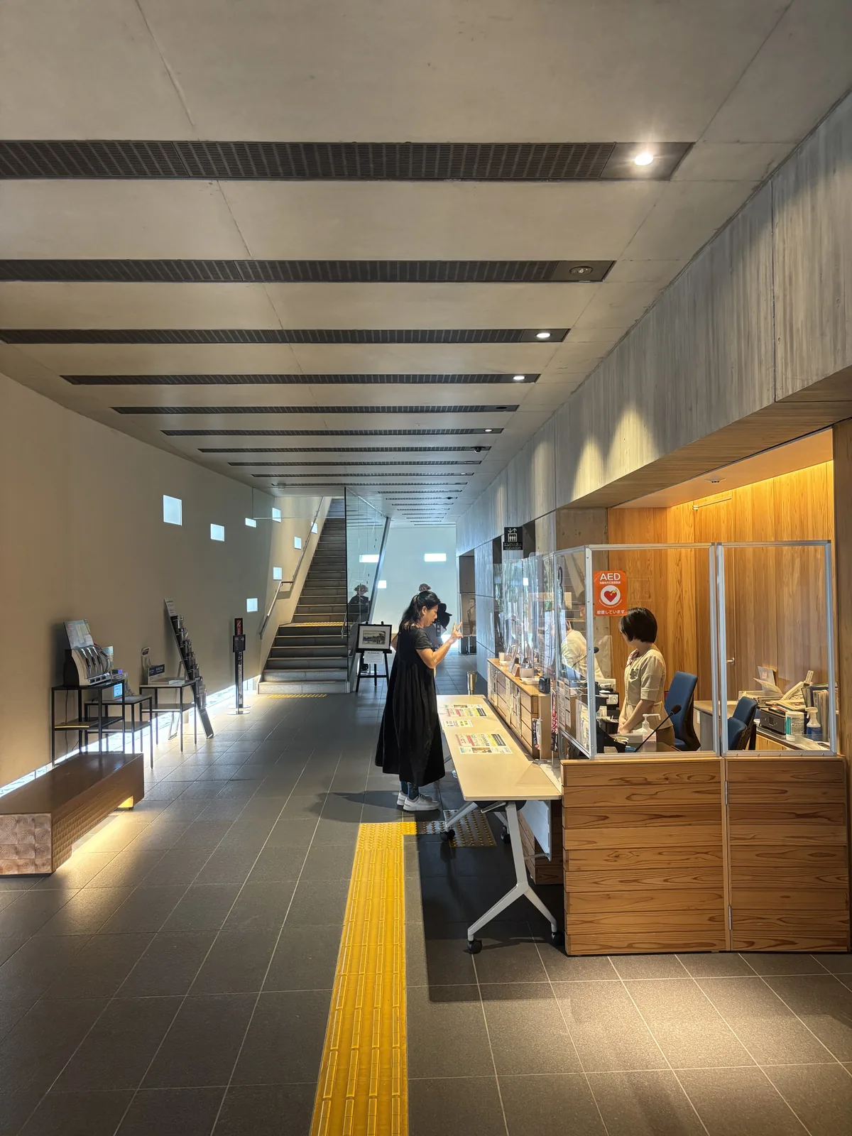 新館一樓的細長大廳，右側是木製的受付櫃台與透明隔板，左側是一道開著零星小方窗的白牆，地面鋪深灰色石材與黃色導盲磚，天花有等距的深色線條，盡端是一道上行的階梯