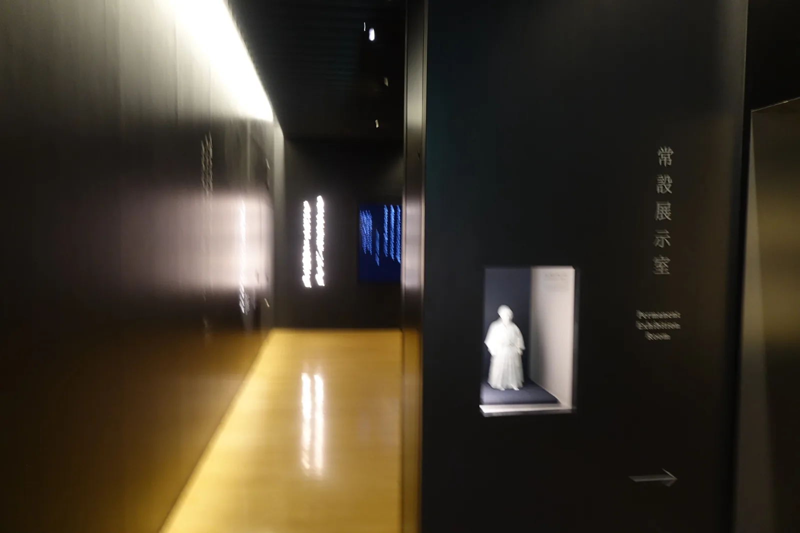 新館二樓常設展示室的入口，深色的牆面上嵌著一個發光的壁龕，龕內立著一尊白色的人像，壁面直書「常設展示室」，地面反射著頂部的線狀照明