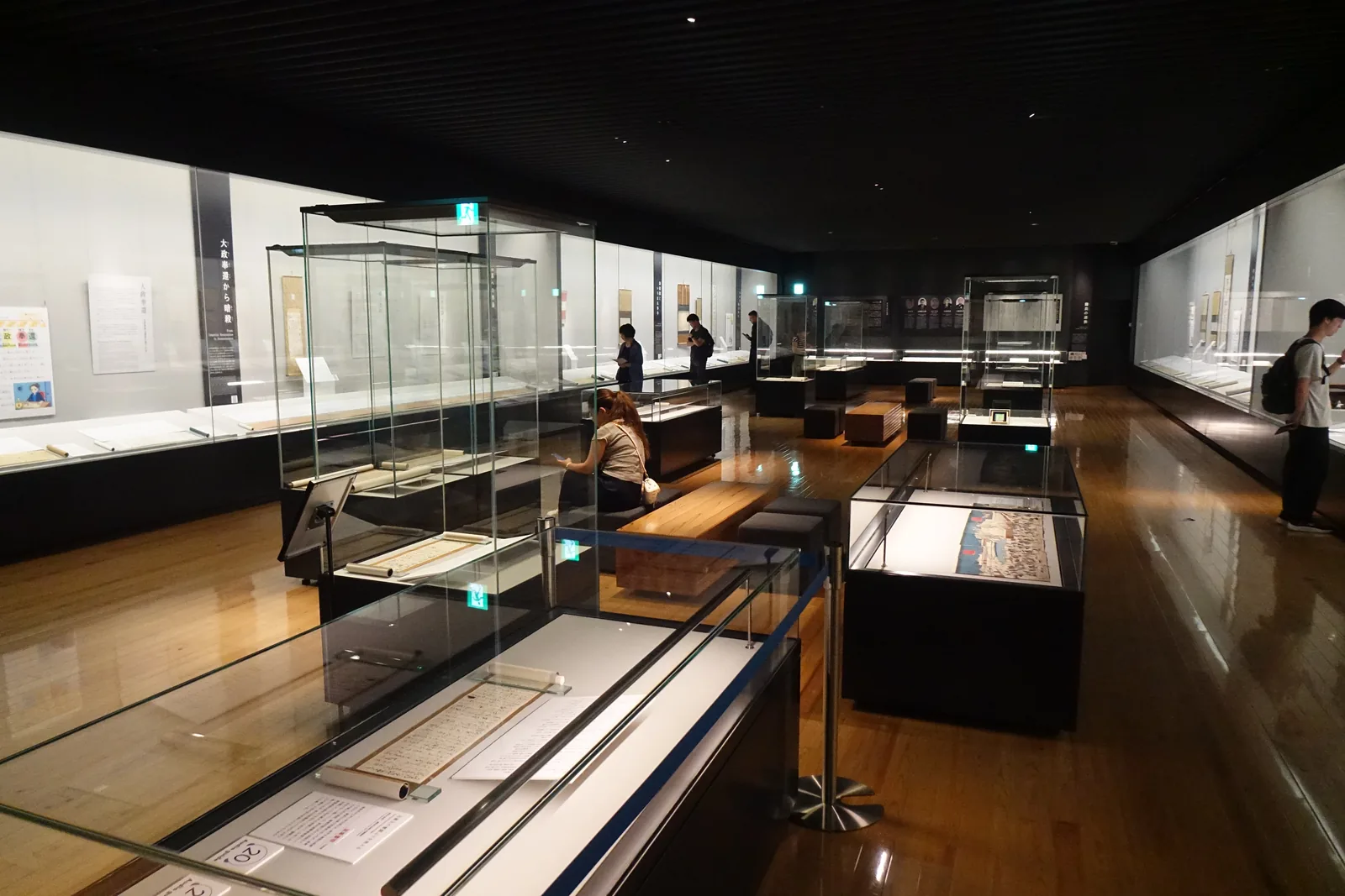 新館二樓常設展示室，木質地板上排列著玻璃的獨立展示櫃，兩側是沿牆的壁面展示櫃，天花是黑色的，參觀者在展示櫃之間移動