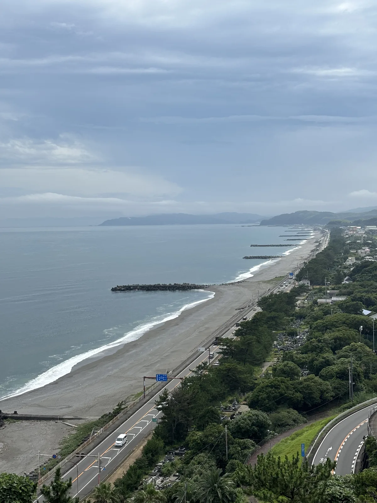自本館屋頂俯瞰浦戶西側的海岸，筆直的沙灘沿海岸線延伸到遠方，沙灘外側是幾道消波塊堤，沙灘與道路之間是茂密的松林，遠處是低平的海岸與山稜