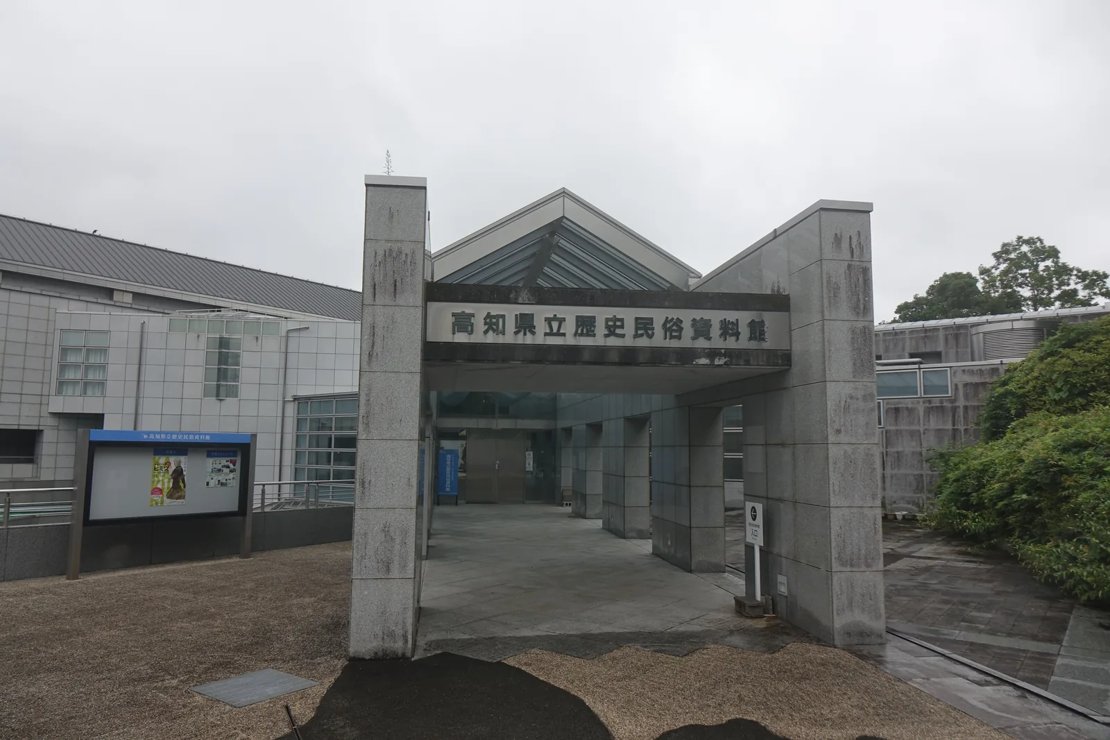 高知縣立歷史民俗資料館的入口門廊，兩根花崗岩方柱撐起刻有館名的橫樑，樑後是斜向的玻璃屋頂，門廊下是通往入口的通道，左側是一座說明看板與館舍的灰色外牆