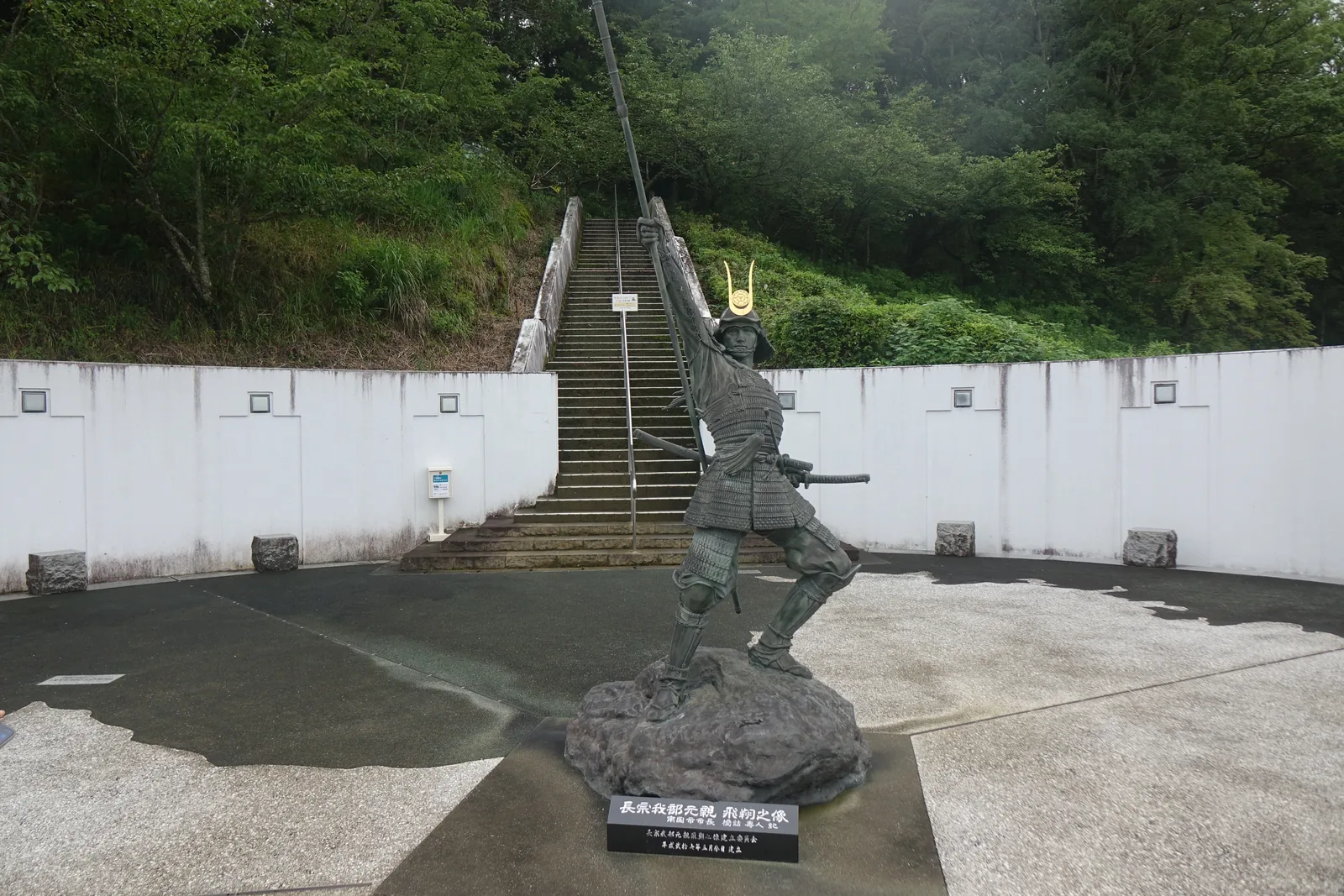 資料館前面廣場上的長宗我部元親像，持槍前傾的銅像立在岩座上，背後是一道白色的擋土牆與一段上山的石階，石階兩側是樹林