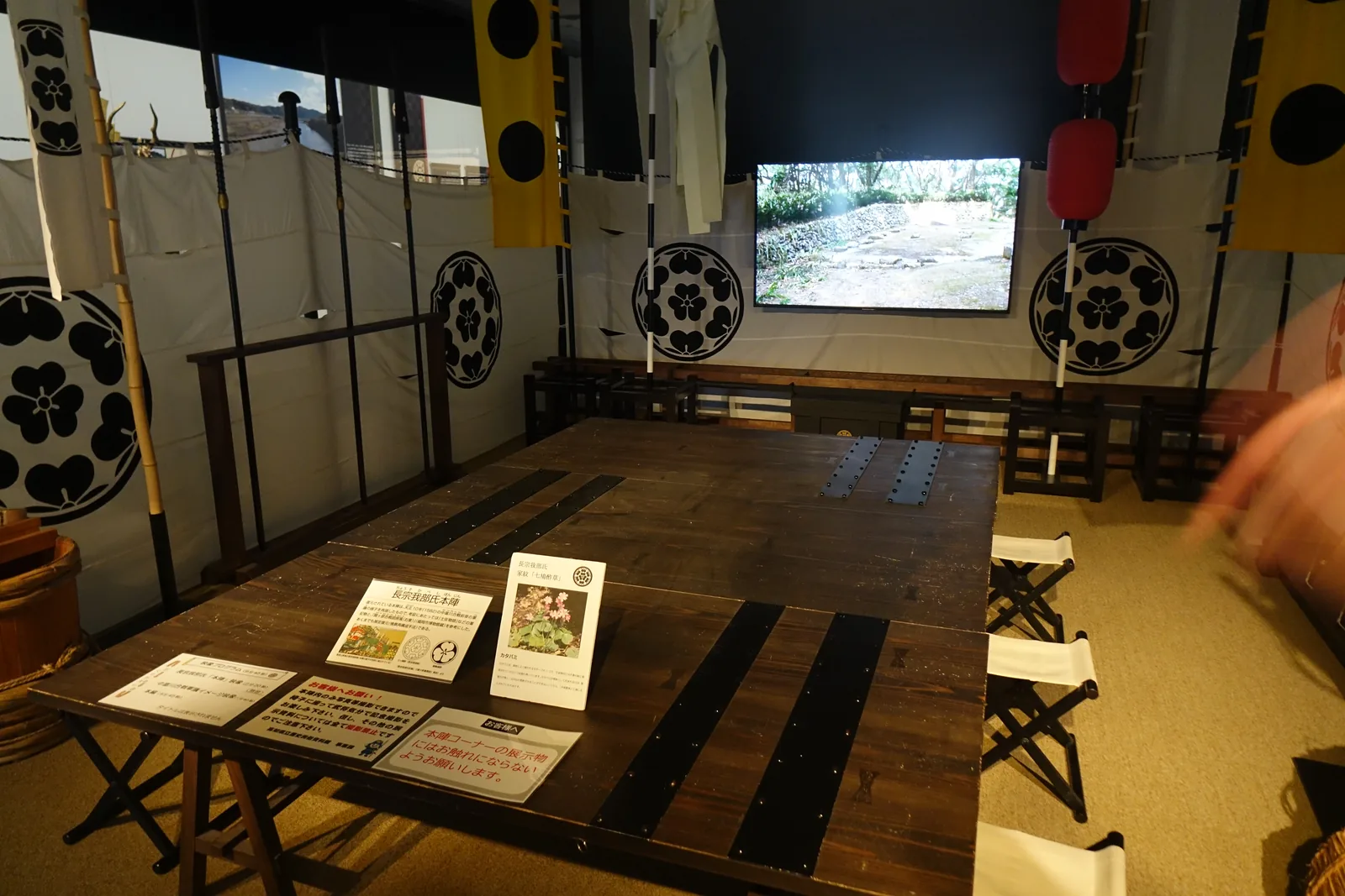 長宗我部展示室中央的本陣再現，白布圍幕上印著黑色的家紋，四周插著黃黑兩色的旗幟，中央是一張深色的長桌與摺椅，後方牆上是播放城跡影像的螢幕