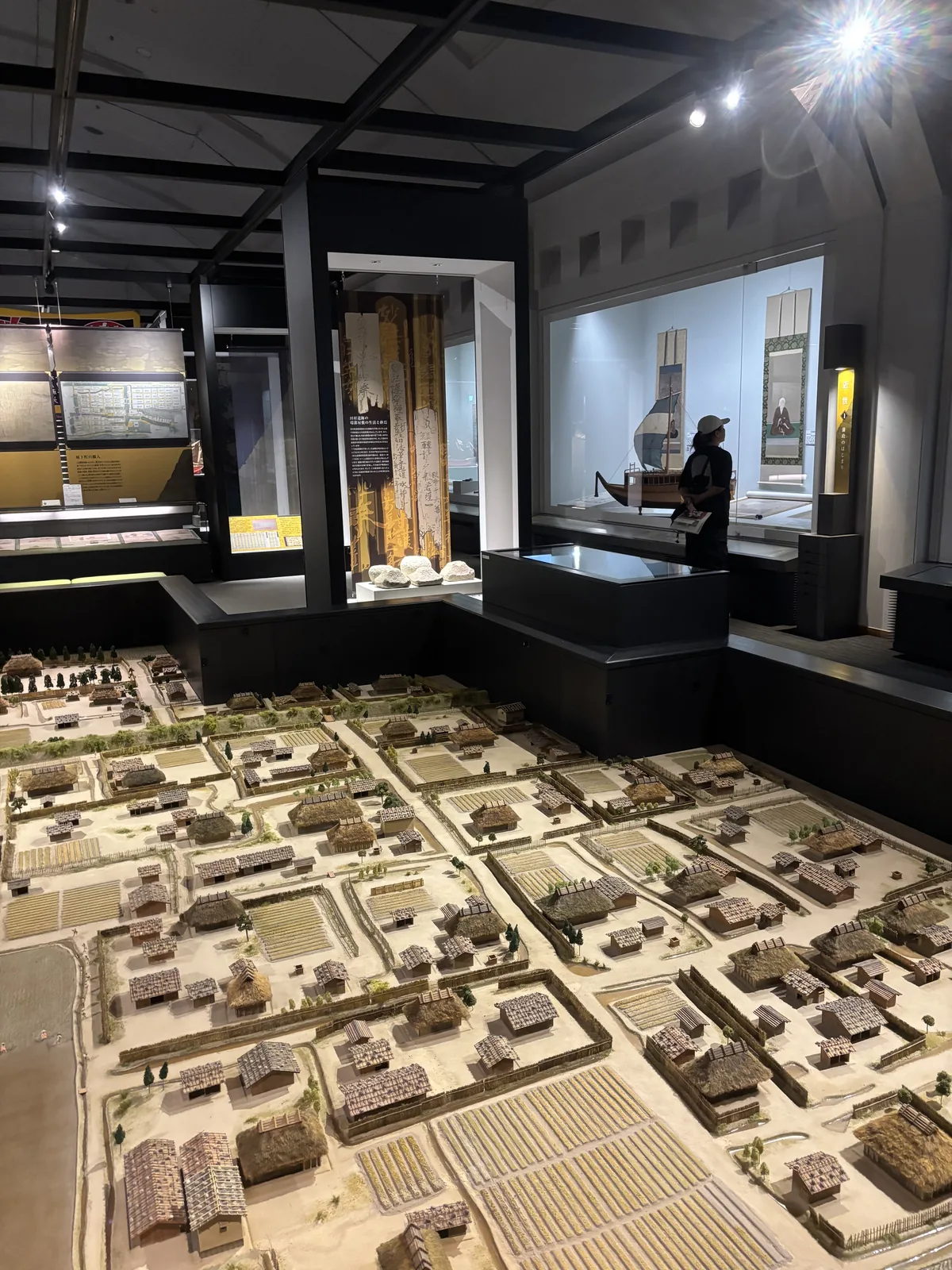 三樓總合展示室，前景是一座低平的大型聚落復原模型，模型之後是黑色框架構成的展示櫃與立面展示，遠處一名參觀者站在展示櫃前
