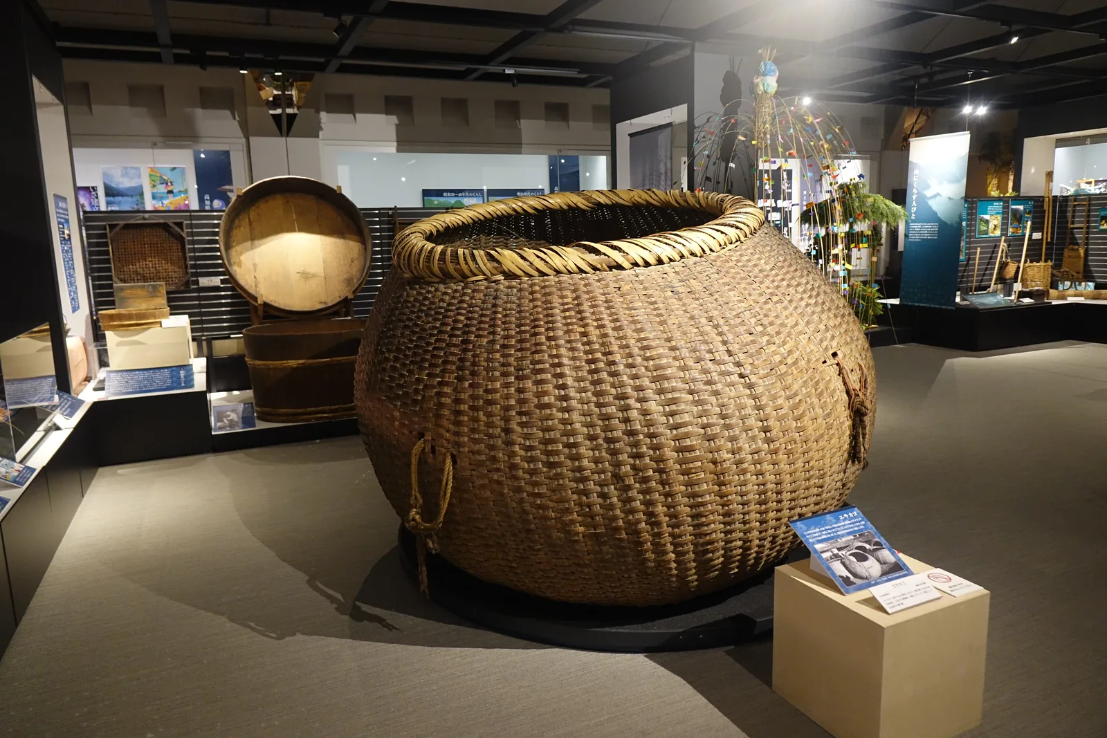 總合展示室中的一個大型竹編籠，置於展示台上，後方是木桶與漁具的展示，遠處是藍色的解說板與天花的格子