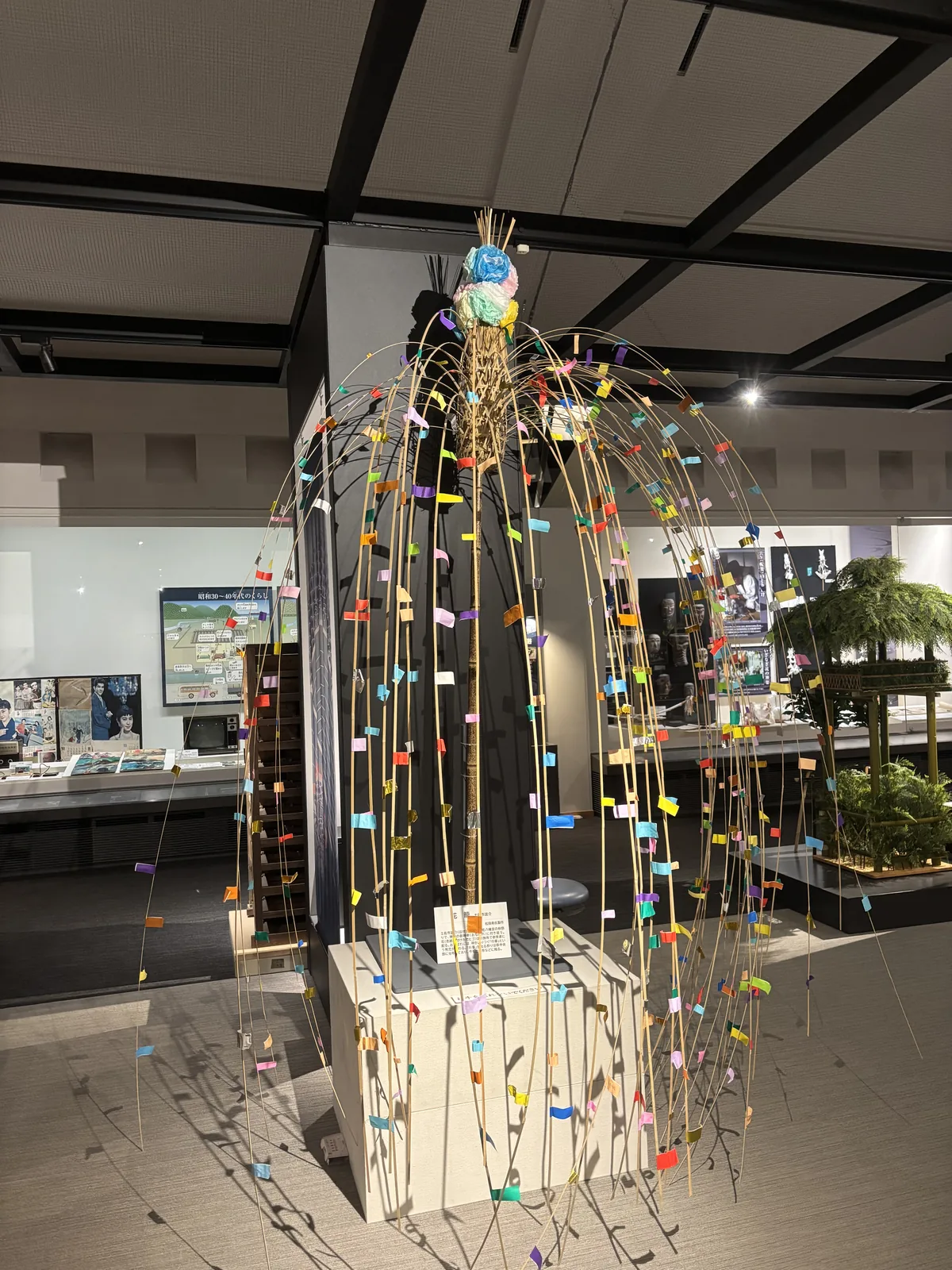 總合展示室中的祭典展示，前景是一株插滿彩色紙條的竹製祭具，後方是白色的展示櫃與另一座綠葉裝飾的水棚模型，天花是深色的格子