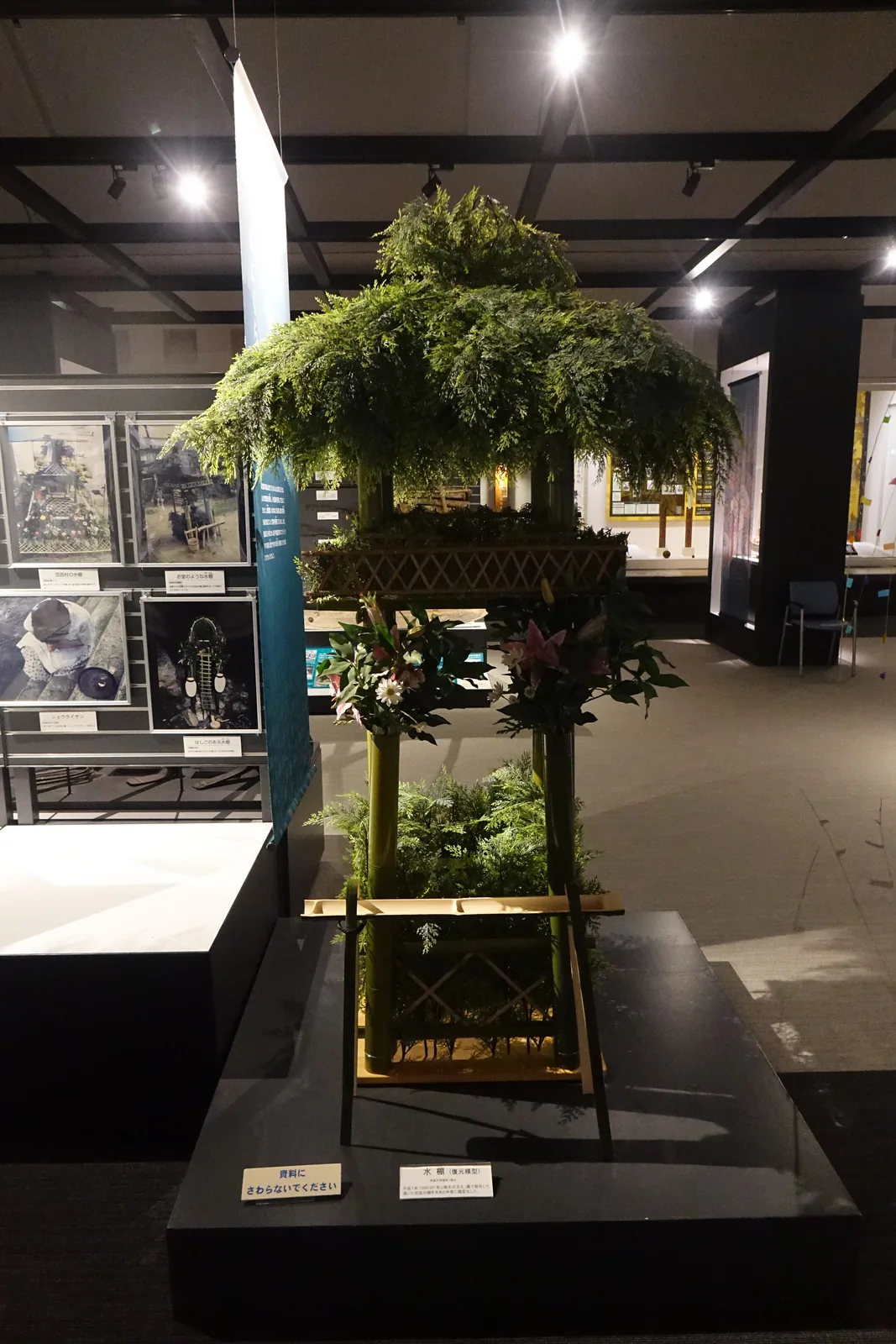 總合展示室中的水棚復原模型，以竹組成的棚架上覆滿杉葉與花，置於黑色的展示台上，左側是盆行事的照片展示