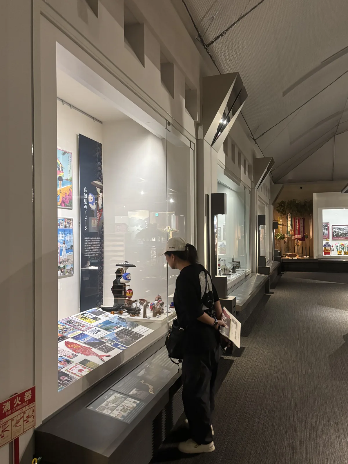 沿外牆的展示櫃廊，左側是整排的玻璃展示櫃與鄉土玩具的展示，天花沿屋頂坡度斜下，一名參觀者在櫃前觀看，廊道盡端連接下一個展示空間