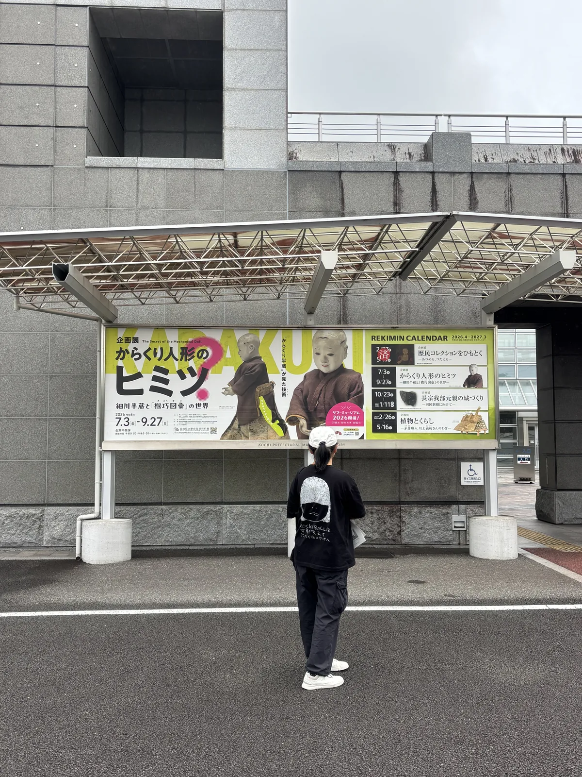 資料館外的鋼構雨庇之下掛著一幅黃綠色的企畫展看板，看板上是機關人偶的照片，一名參觀者站在看板前，背後是灰色的館舍外牆