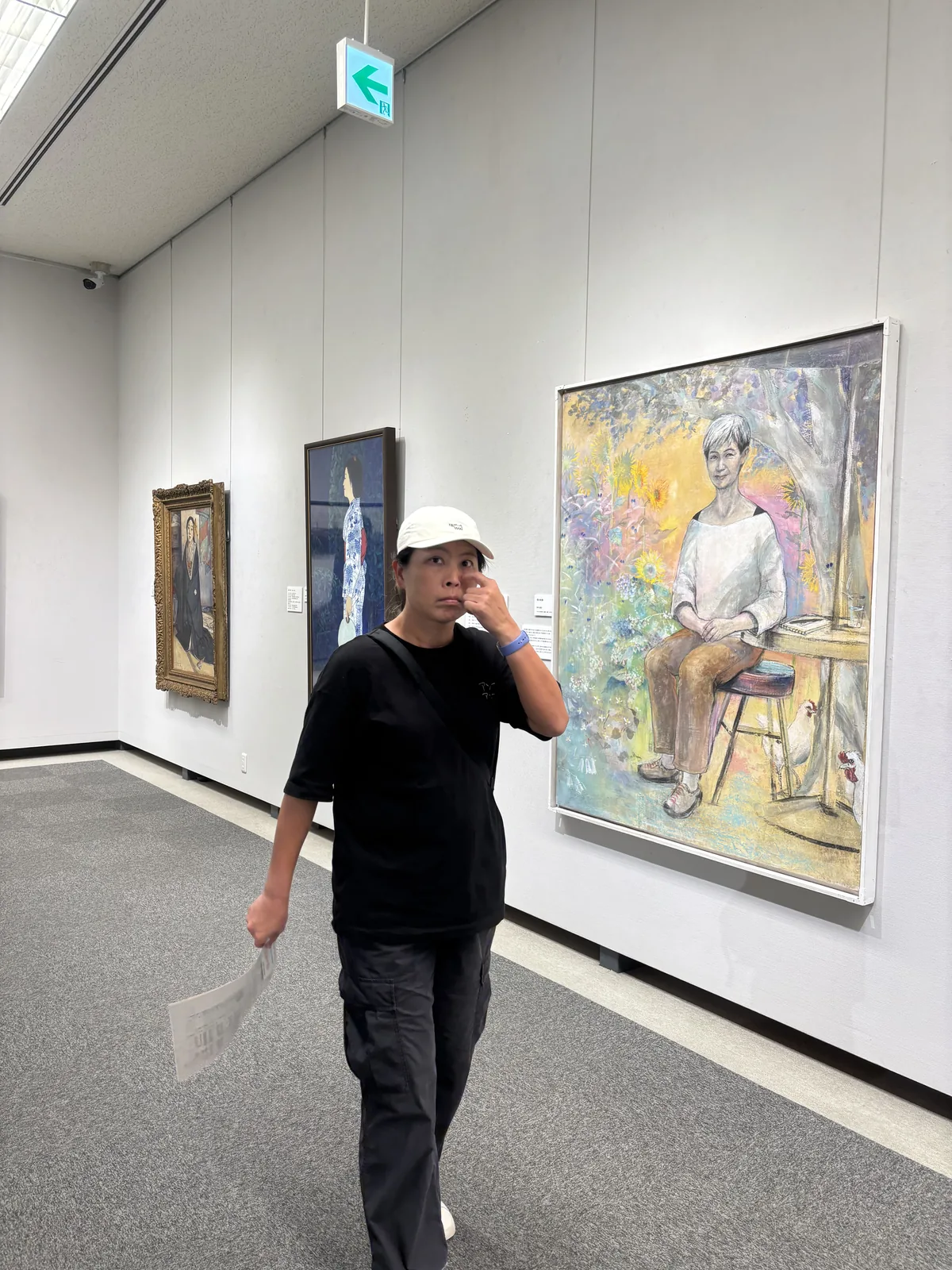香美市立美術館的展示室，灰色地毯上一名參觀者走過，右側白牆上掛著一幅人物油畫，遠處牆上是另外兩幅畫作，牆面頂端與天花之間是一條光帶，左上方是綠色的出口標示
